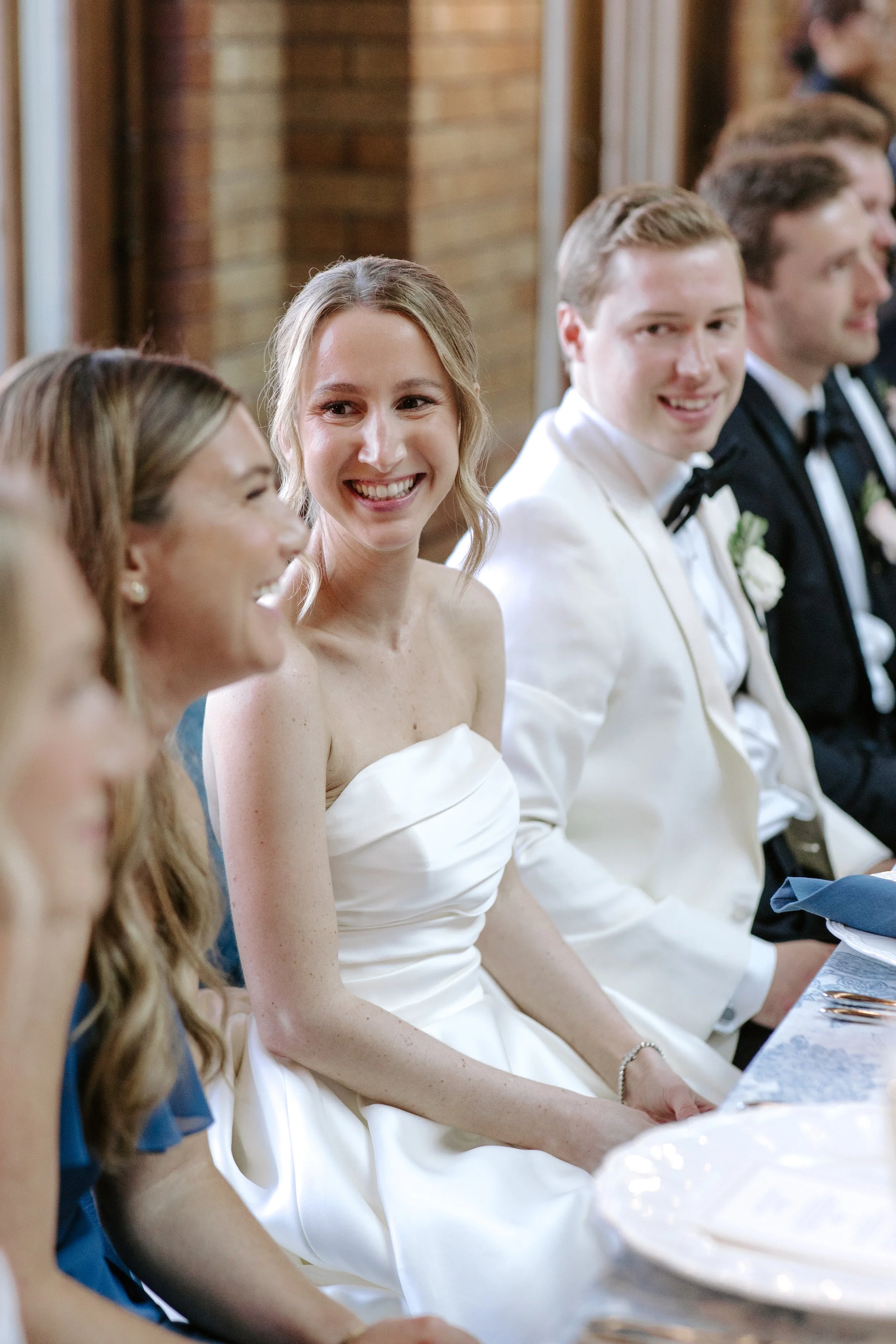 Group of people sitting at a banquet table, with a woman in a white dress smiling, likely at a wedding or formal event.