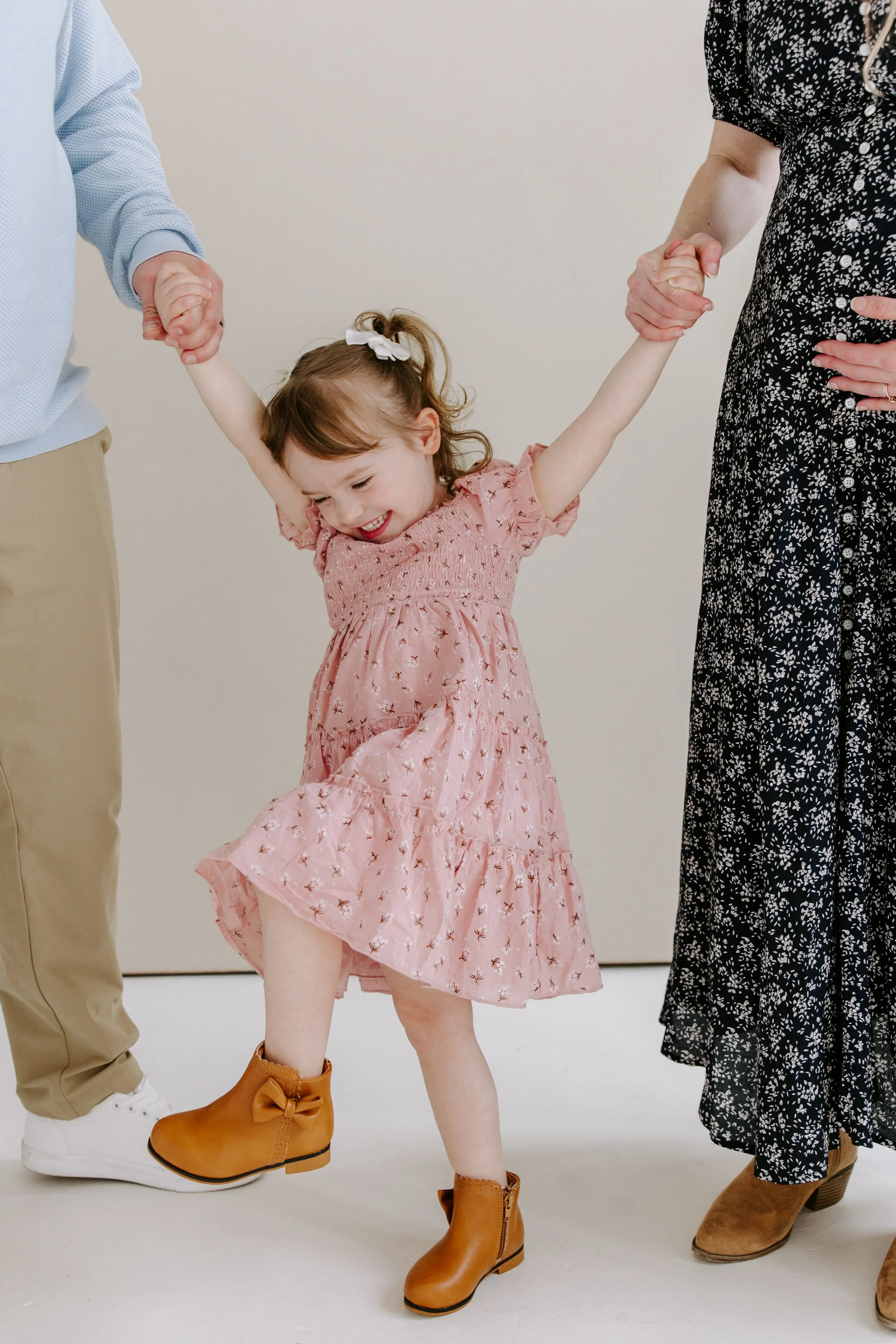 A young girl in a pink dress and brown boots is holding hands with two adults, one in beige pants and white sneakers, and the other in a black floral dress and brown boots, as they dance or walk together against a plain beige background.