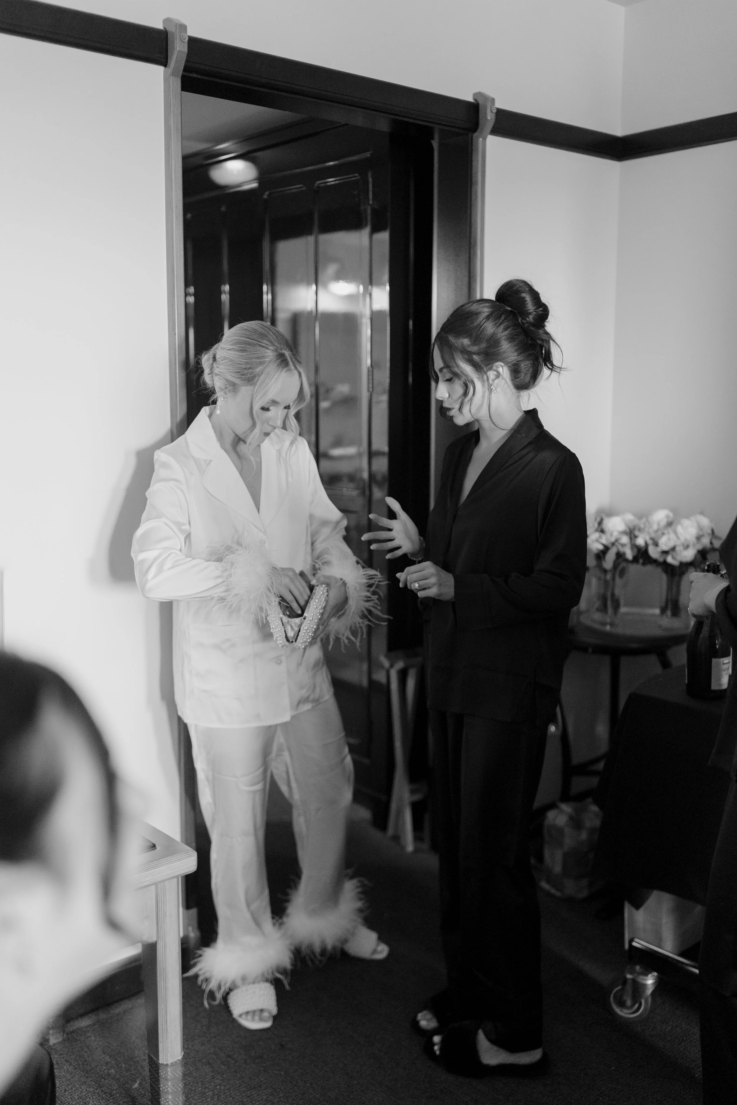 Two women, one in a white outfit with feather details and the other in a dark blazer, are standing in a room. The woman in white is looking down at an item, while the woman in dark is gesturing with her hand. There is a flower arrangement on a table 