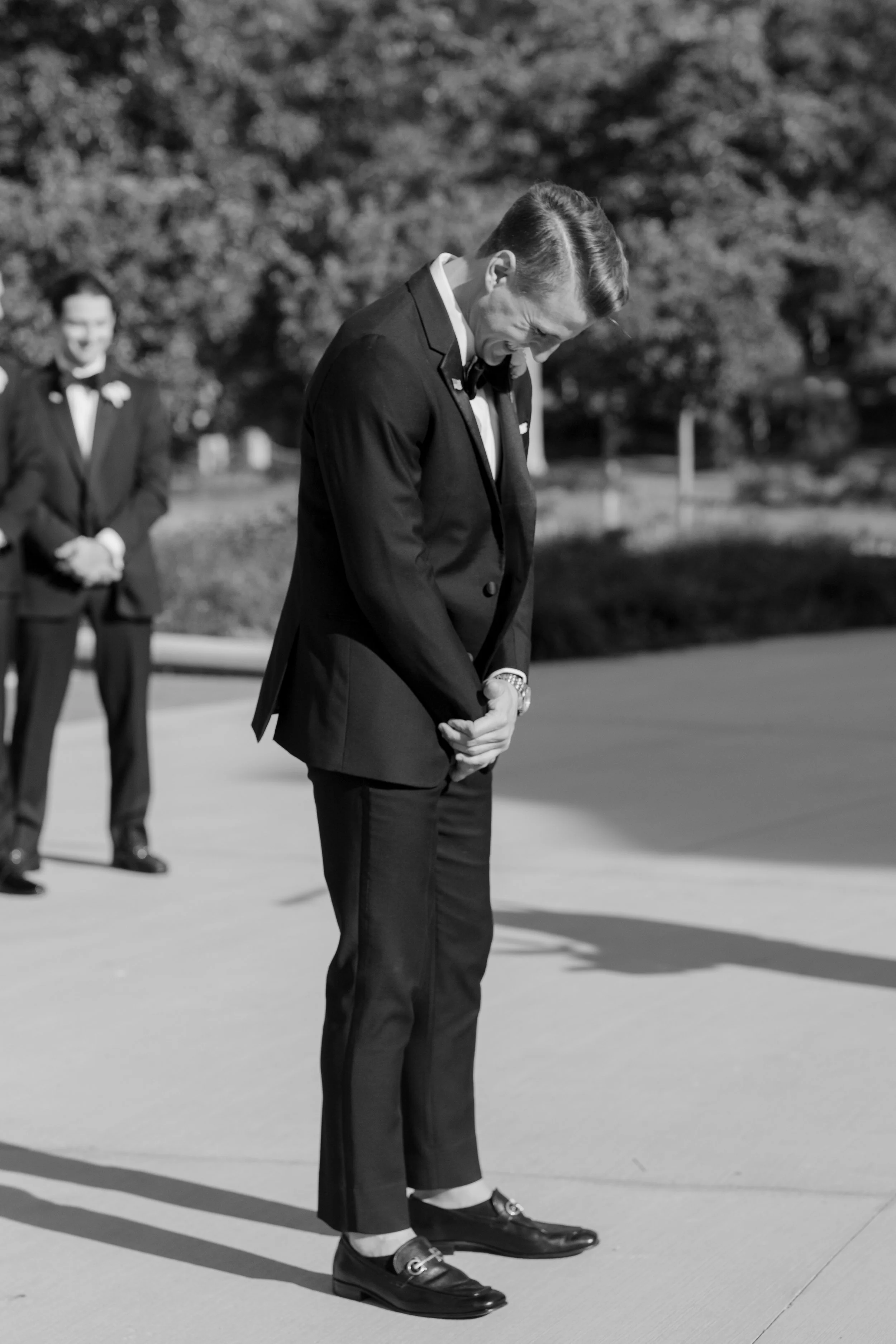 Black and white photo of a man in a tuxedo bowing with his head down, standing on a sidewalk with trees in the background. Another man in a tuxedo is visible in the background, smiling.