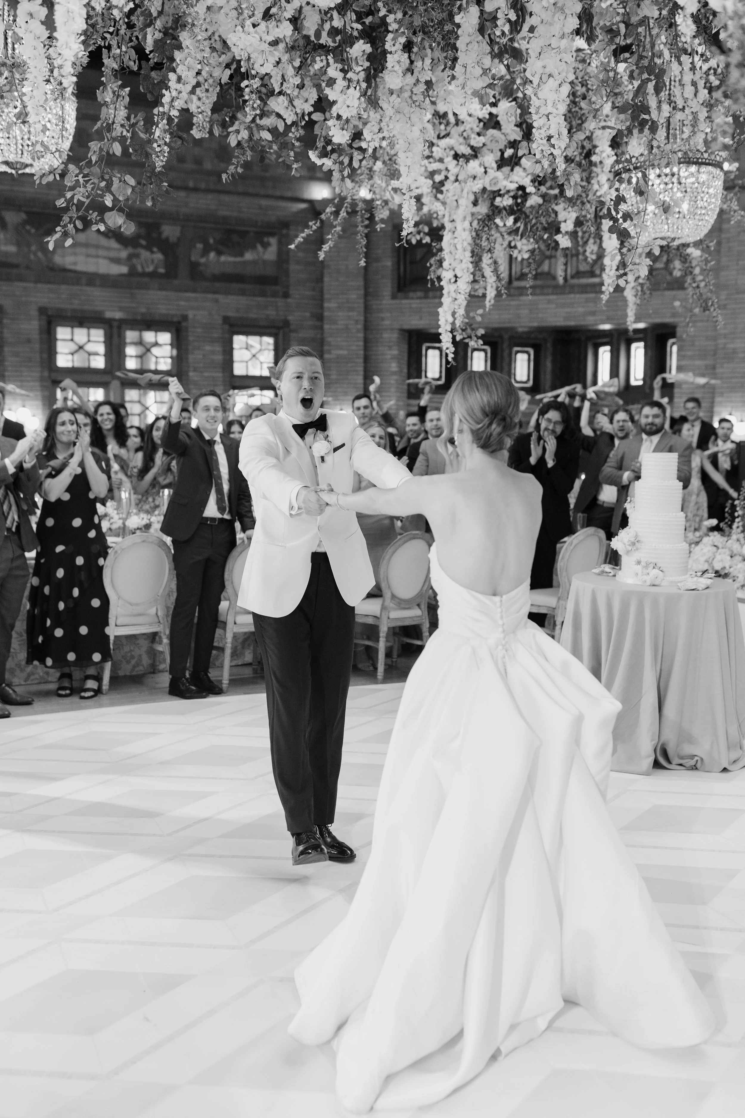 Black and white photograph of a wedding reception with a groom in a tuxedo dancing with a bride in a wedding gown, surrounded by guests, some of whom are raising their arms and clapping, in a decorated hall with hanging floral arrangements and a larg