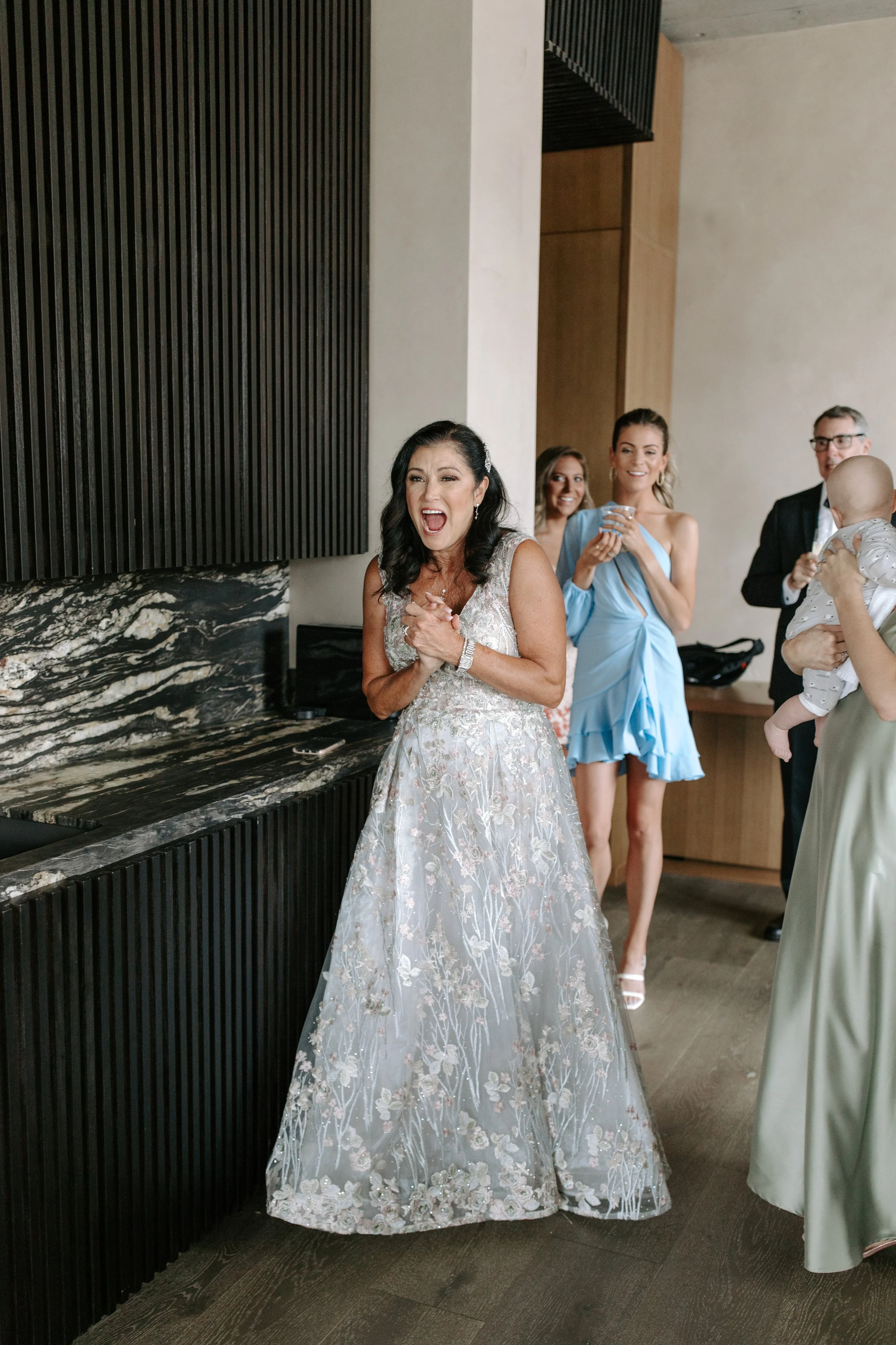 A woman in an elegant wedding gown is smiling and clasping her hands, surrounded by friends and family at a celebration in a modern indoor setting.