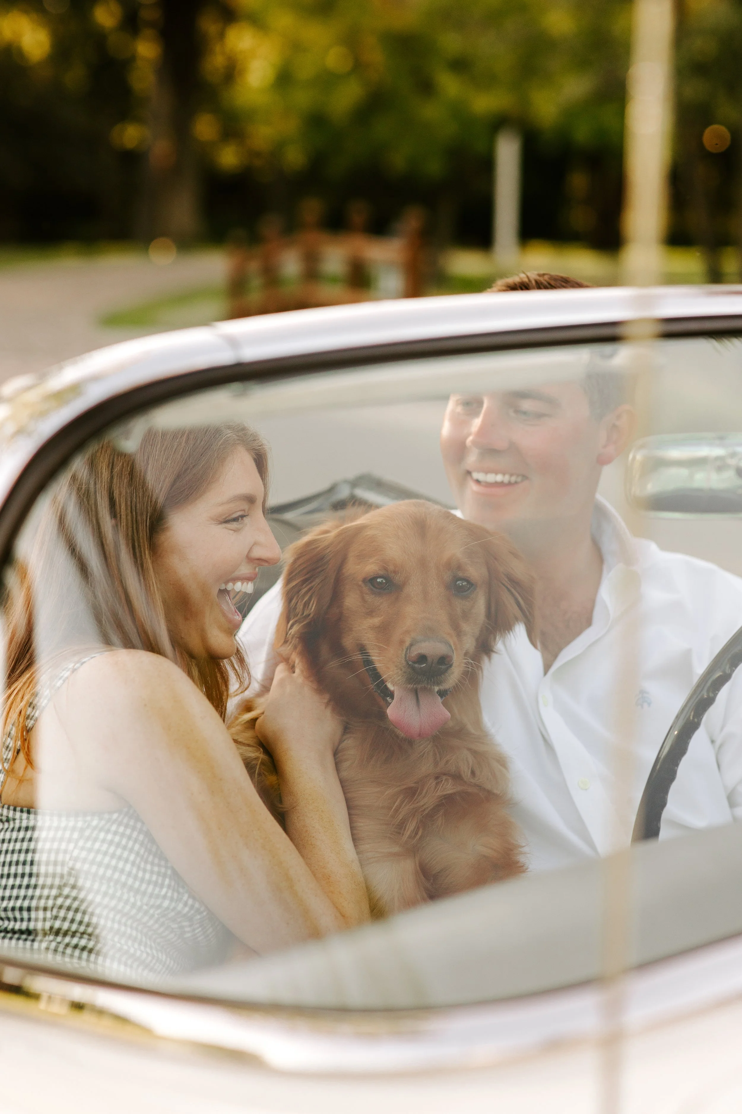 A smiling woman, a smiling man, and a golden retriever dog inside a convertible car during daytime. The woman and man are looking at each other and appear happy. The background features trees and a fence.