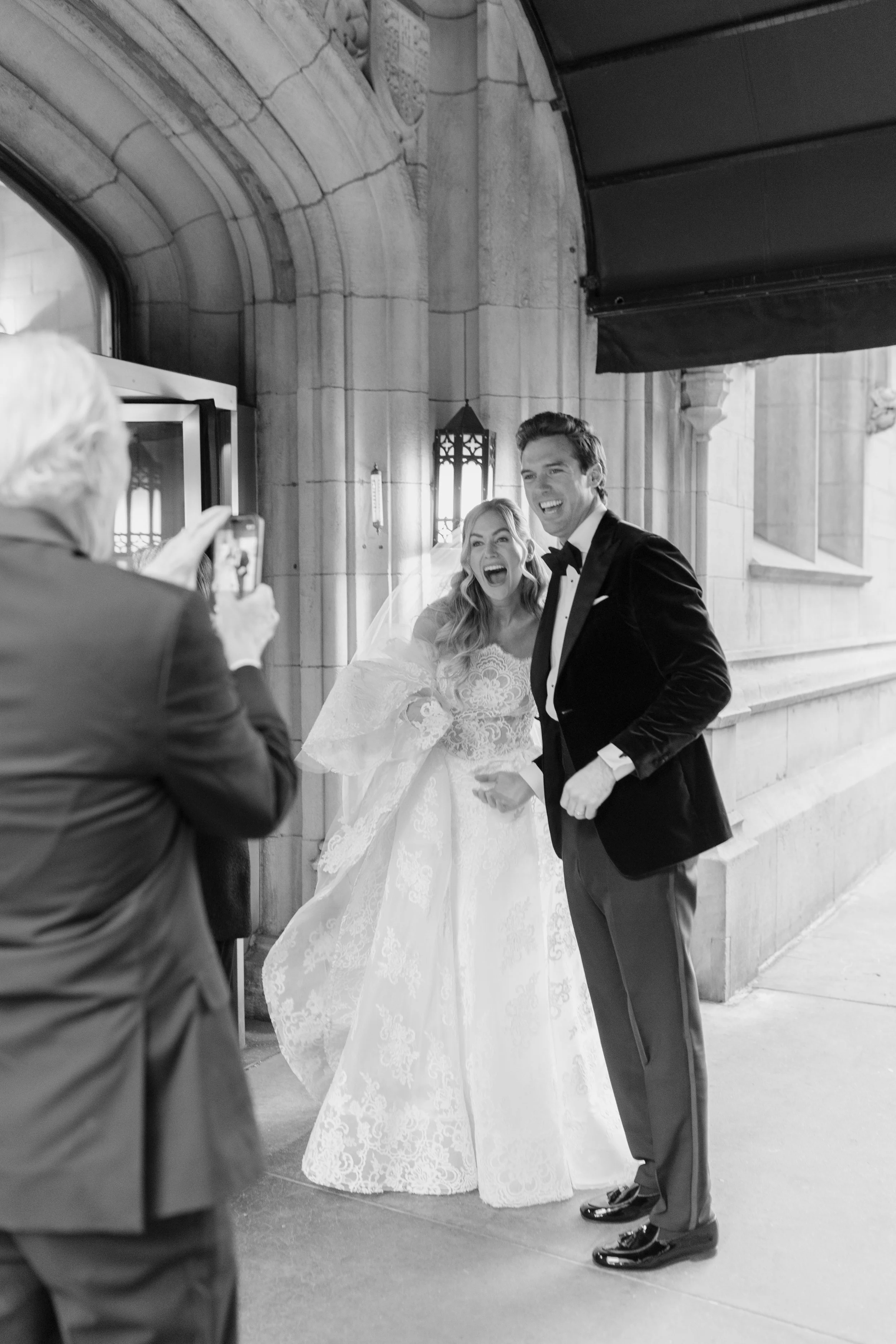A bride and groom smiling and laughing, taking a photo at their wedding outside a stone building, with a person in the foreground holding a smartphone.