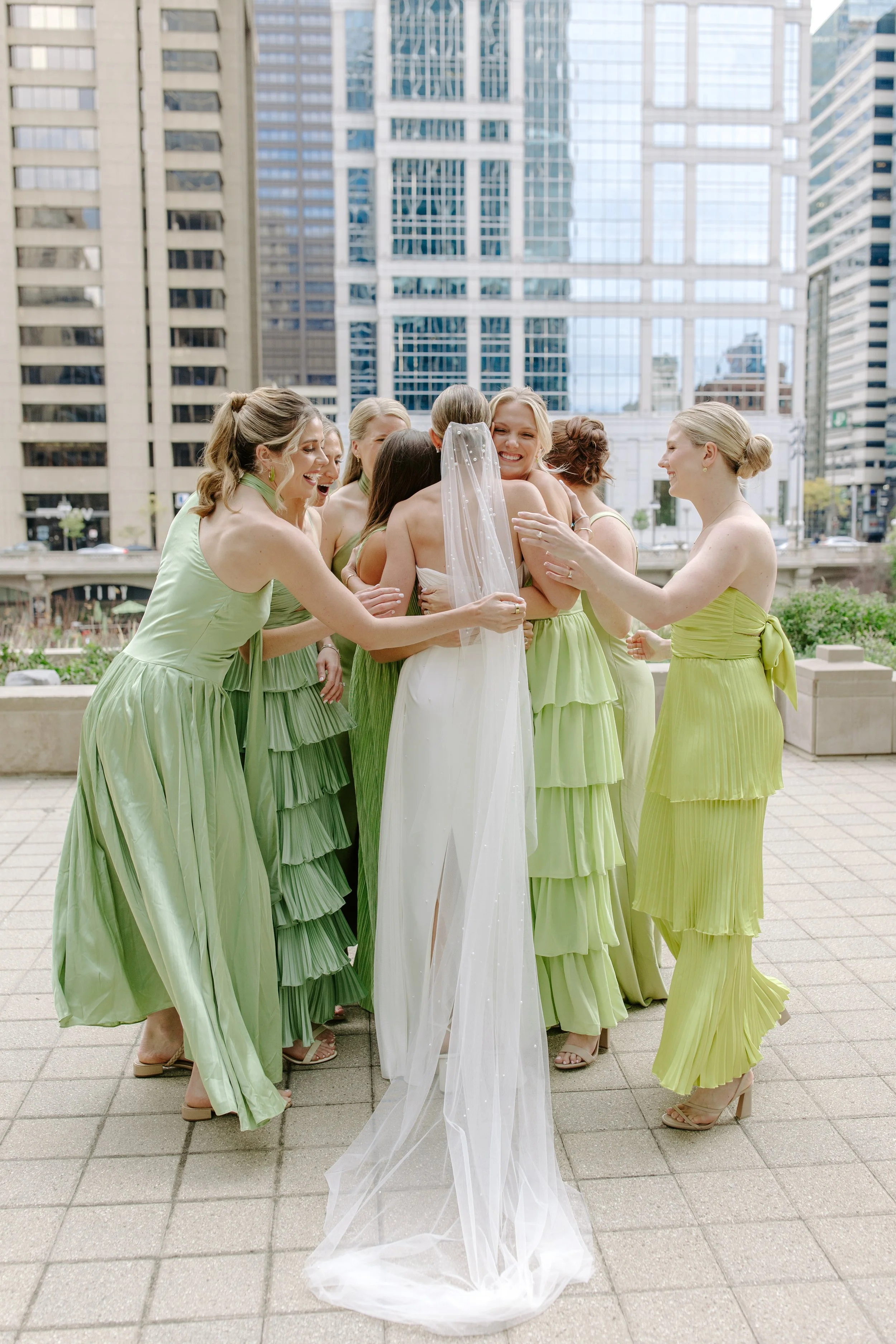 A bride and bridesmaids sharing an embrace outdoors in an urban setting with tall glass buildings in the background. The bridesmaids are dressed in matching light green dresses, and the bride in a white gown with a veil.