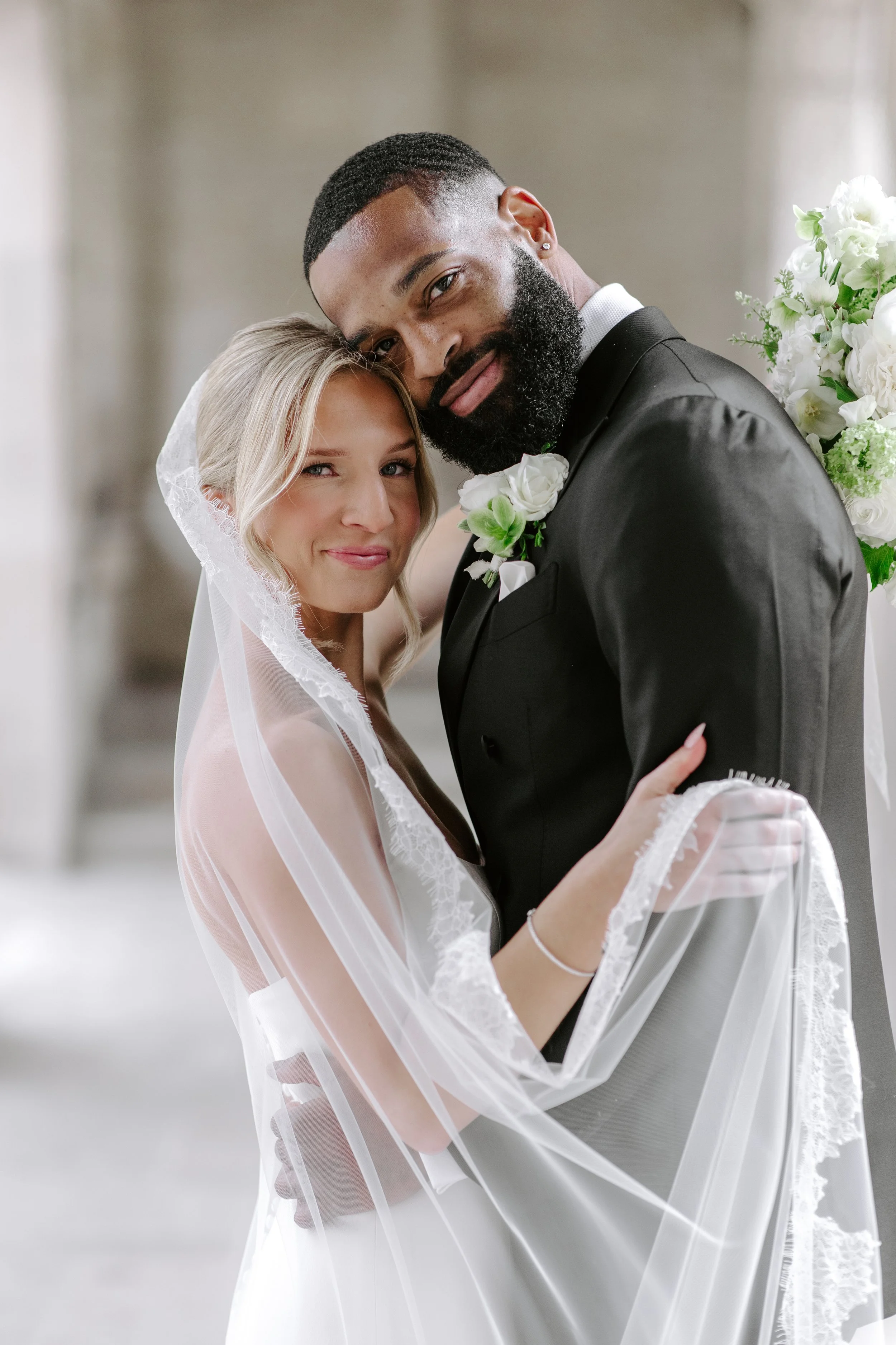 A bride and groom pose closely together during their wedding, smiling and embracing. The bride wears a veil and sheer wedding gown, and the groom wears a black tuxedo with a white flower boutonniere. They are indoors with a plain background.