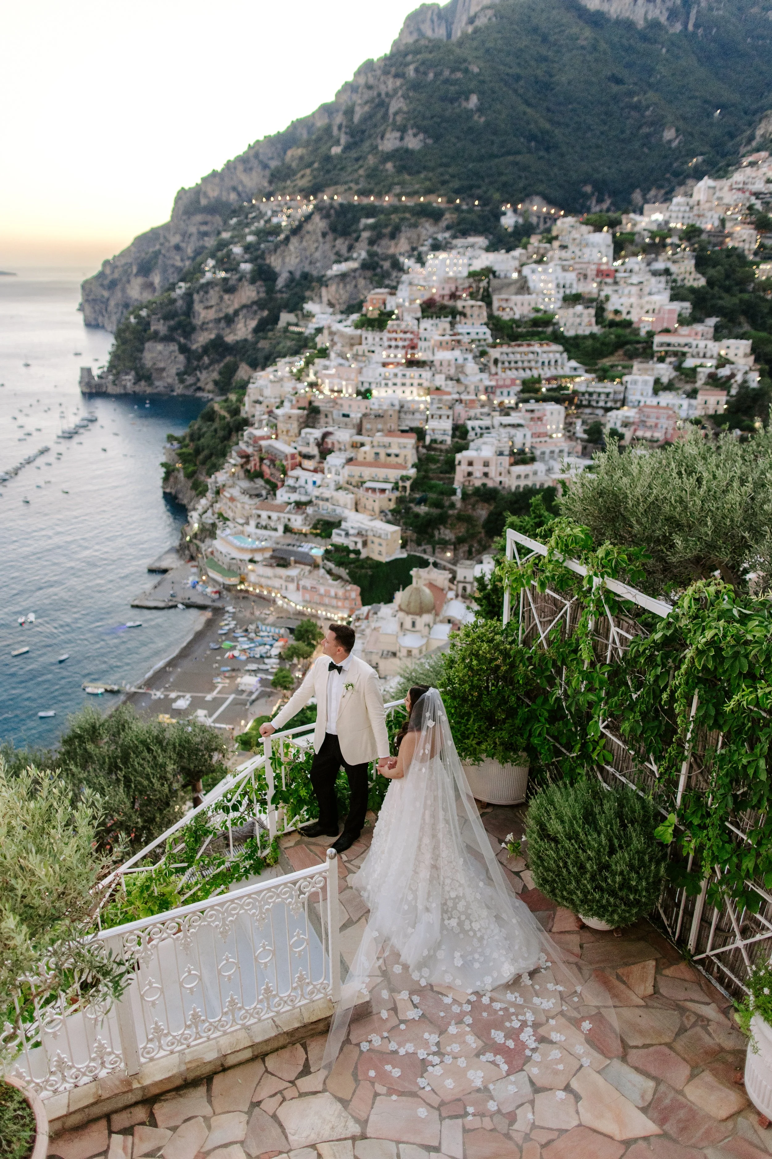 Bride and groom holding hands on a wedding staircase overlooking a coastal town with white buildings, a marina, and a mountain in the background at sunset.