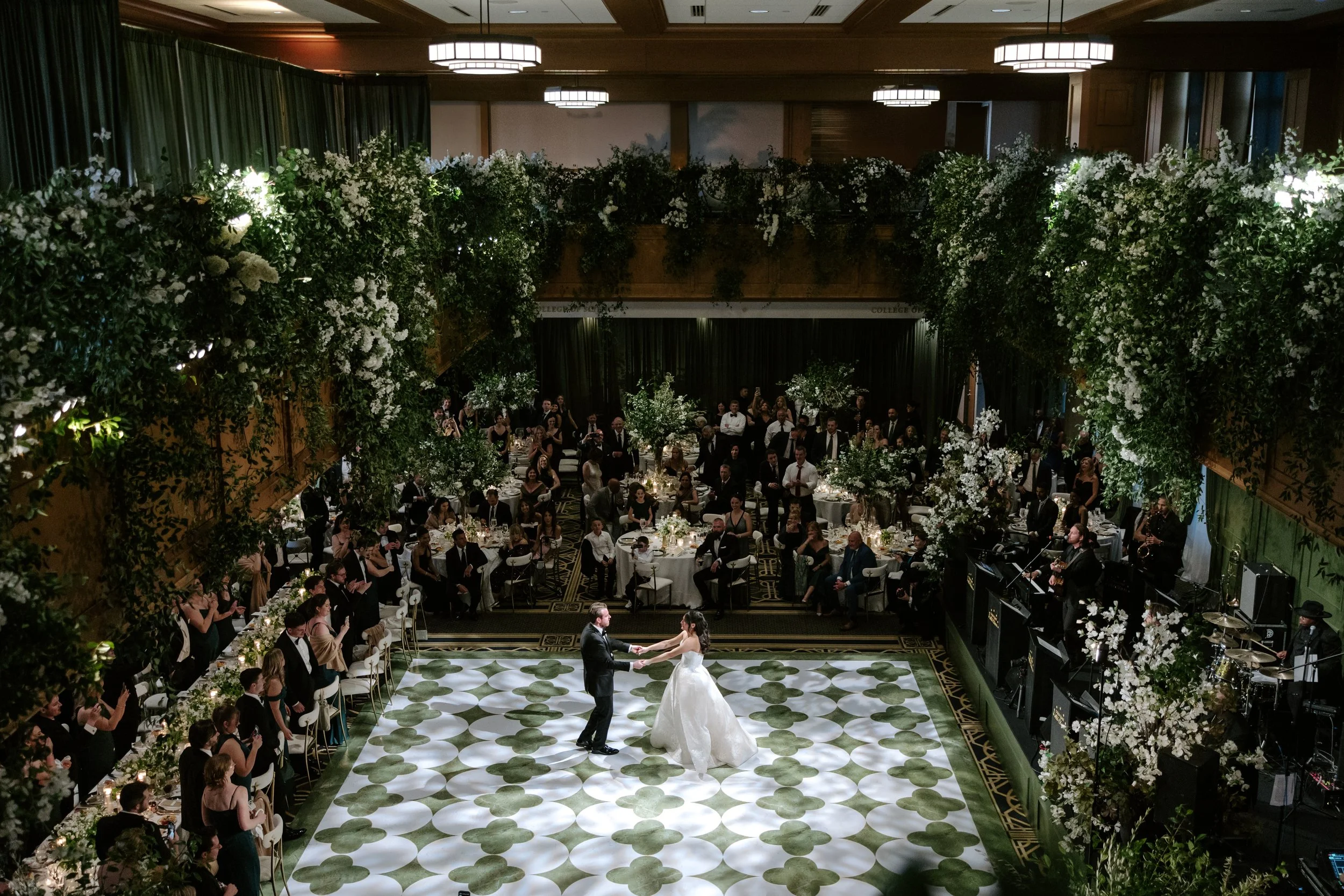 A large wedding reception hall decorated with lush white and green floral arrangements hanging from the ceiling. The bride and groom are dancing on a patterned dance floor at the center of the room, surrounded by seated guests and a band performing l
