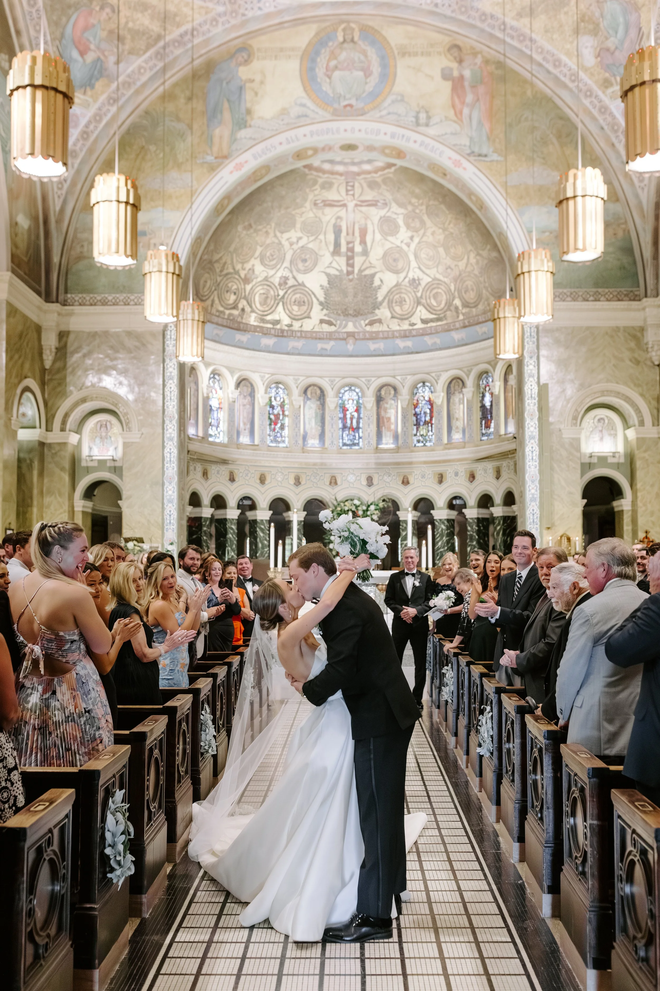 A wedding ceremony inside a church with a couple kissing at the altar, surrounded by friends and family. The church has ornate decorations, stained glass windows, and religious artwork on the ceilings.