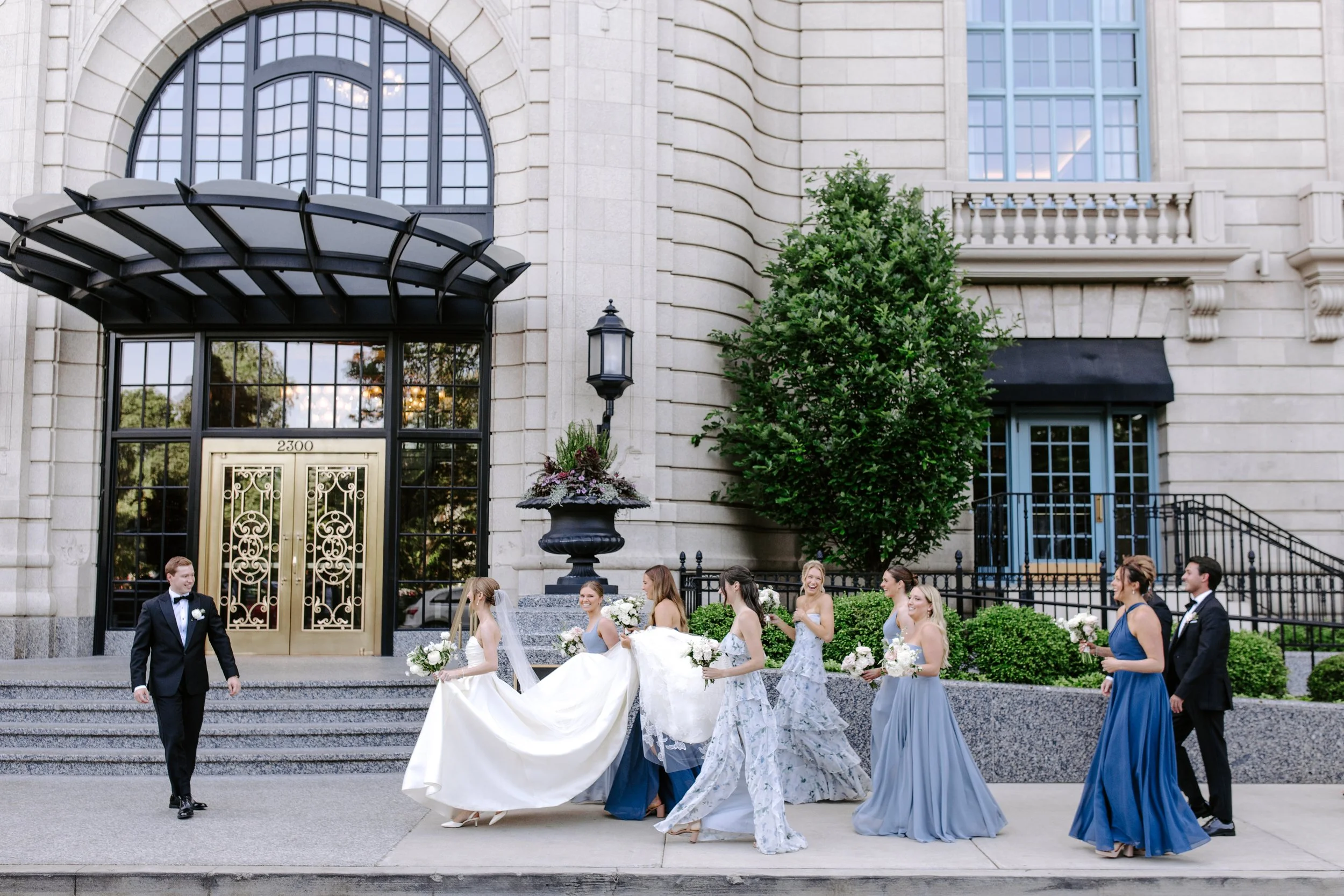 Group of bridesmaids in blue dresses and a bride in a white gown walking down the stairs in front of a large building with arched windows and a decorative black awning, while a groom in a black tuxedo walks alongside them. The scene appears to be a w