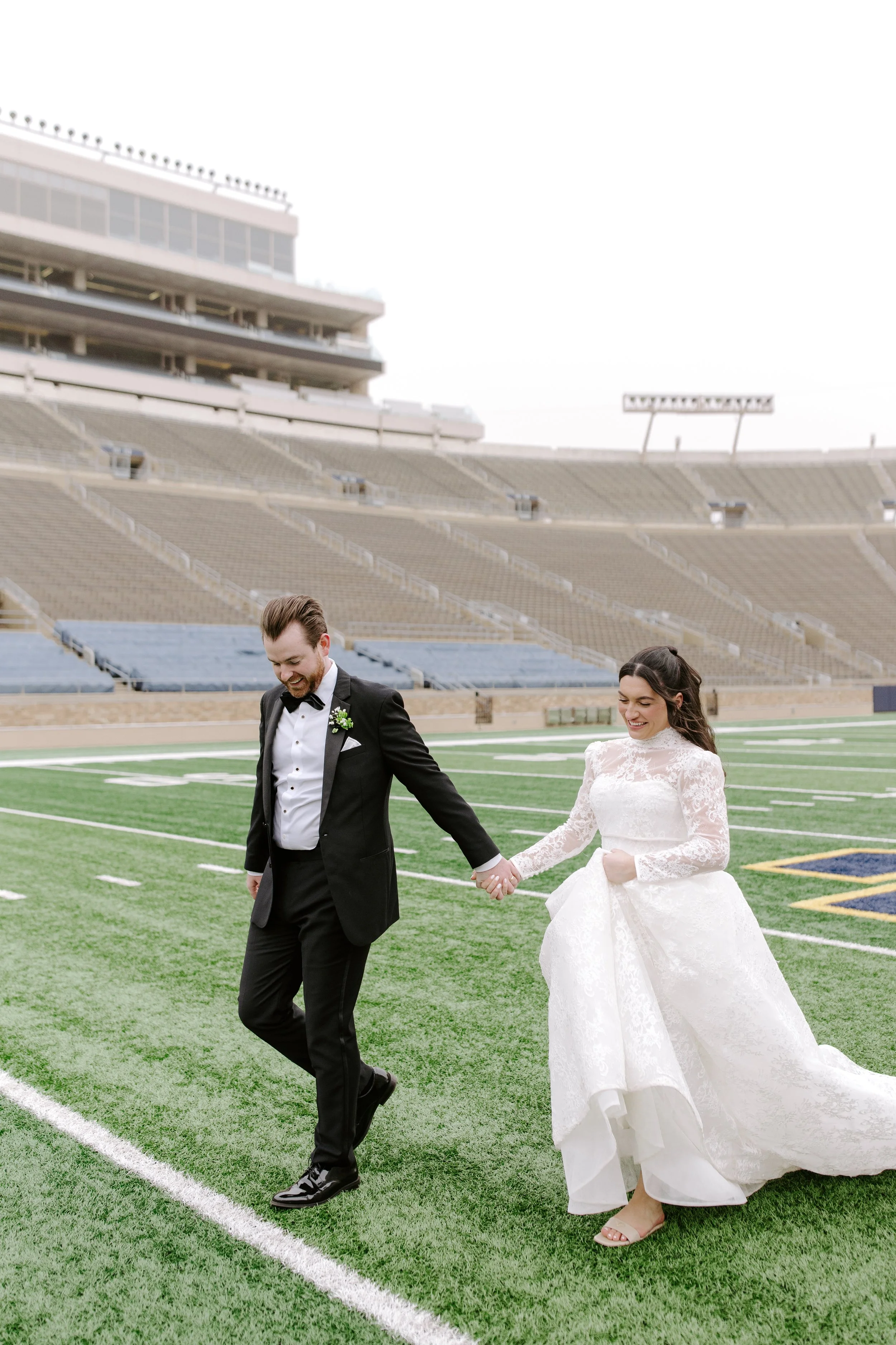 A bride and groom walking hand-in-hand across a football field in a stadium, dressed in wedding attire, with empty seats in the background.