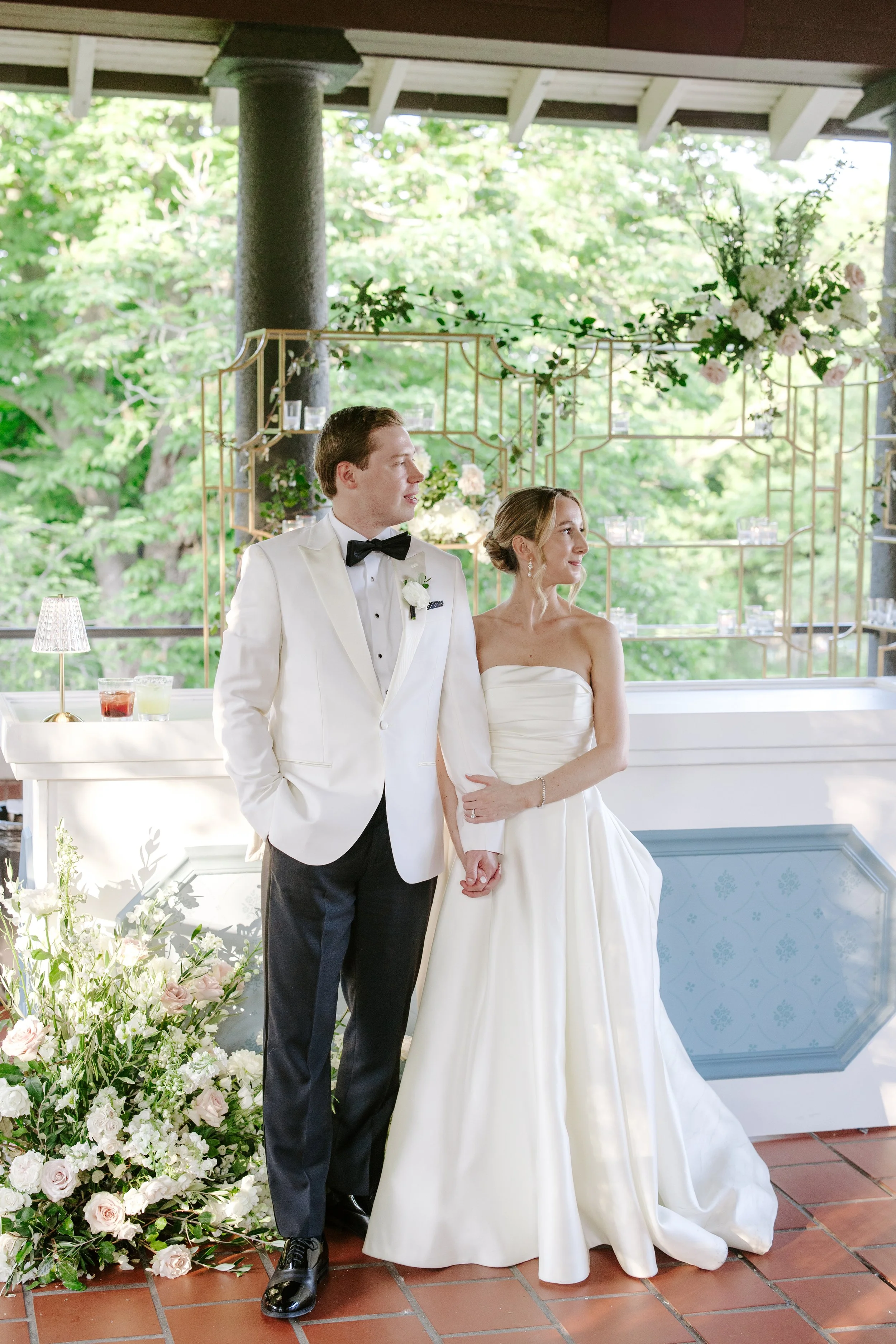 A bride and groom standing hand-in-hand in a wedding reception area decorated with flowers and greenery. The groom is wearing a white tuxedo and the bride is in a strapless white wedding gown.