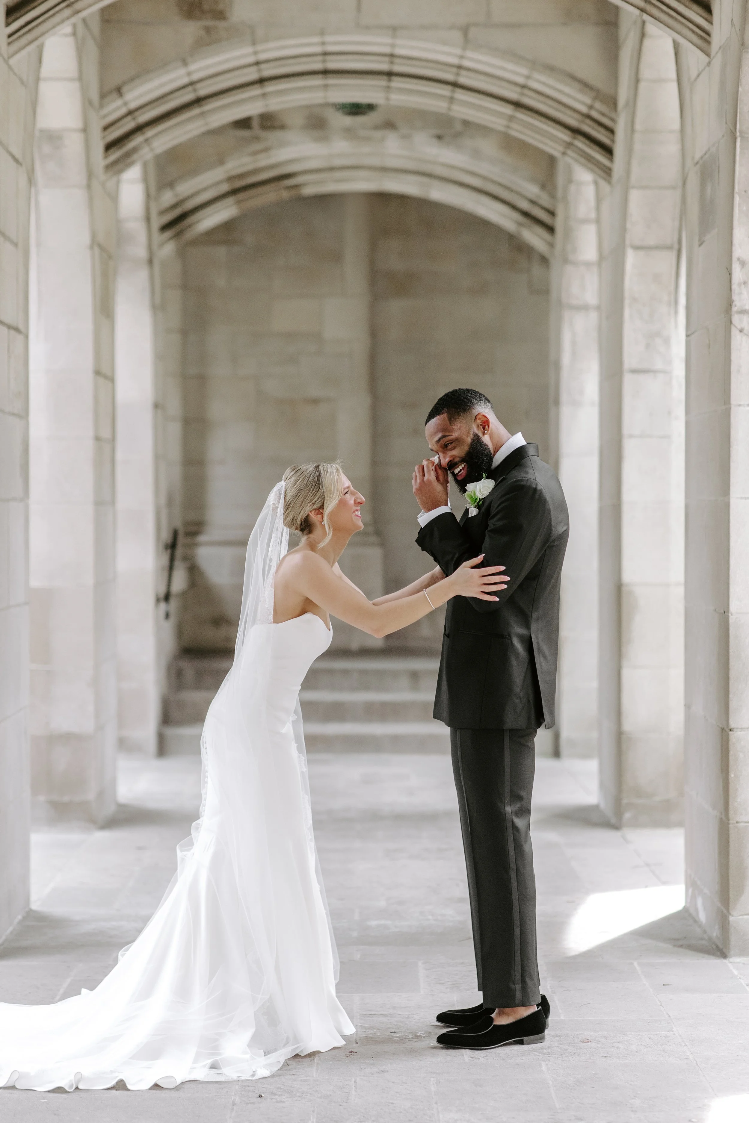 A bride and groom stand together on a stone staircase in a stone church, smiling and looking at each other during their wedding ceremony. The bride is dressed in a white gown with a long train and a veil, and the groom is in a black tuxedo with a whi