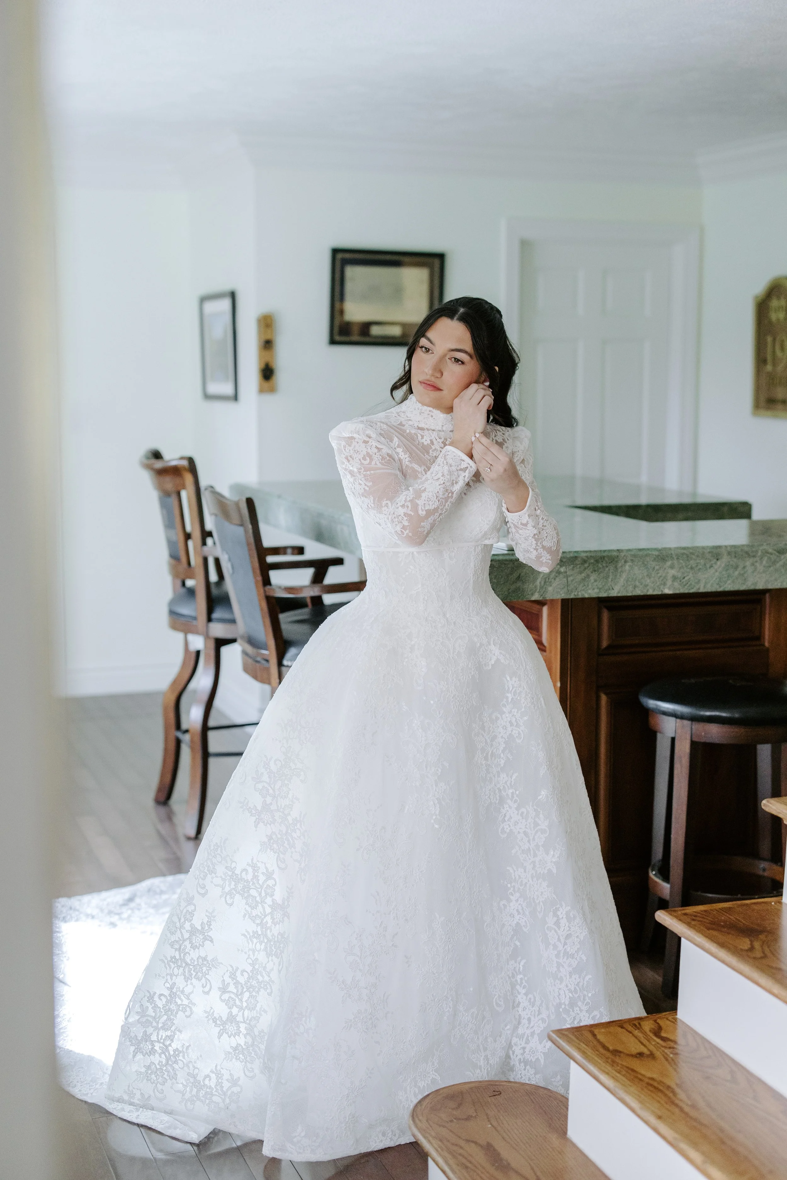 A woman in a white lace wedding dress adjusting her earring inside a home with wooden chairs and framed pictures on the wall.