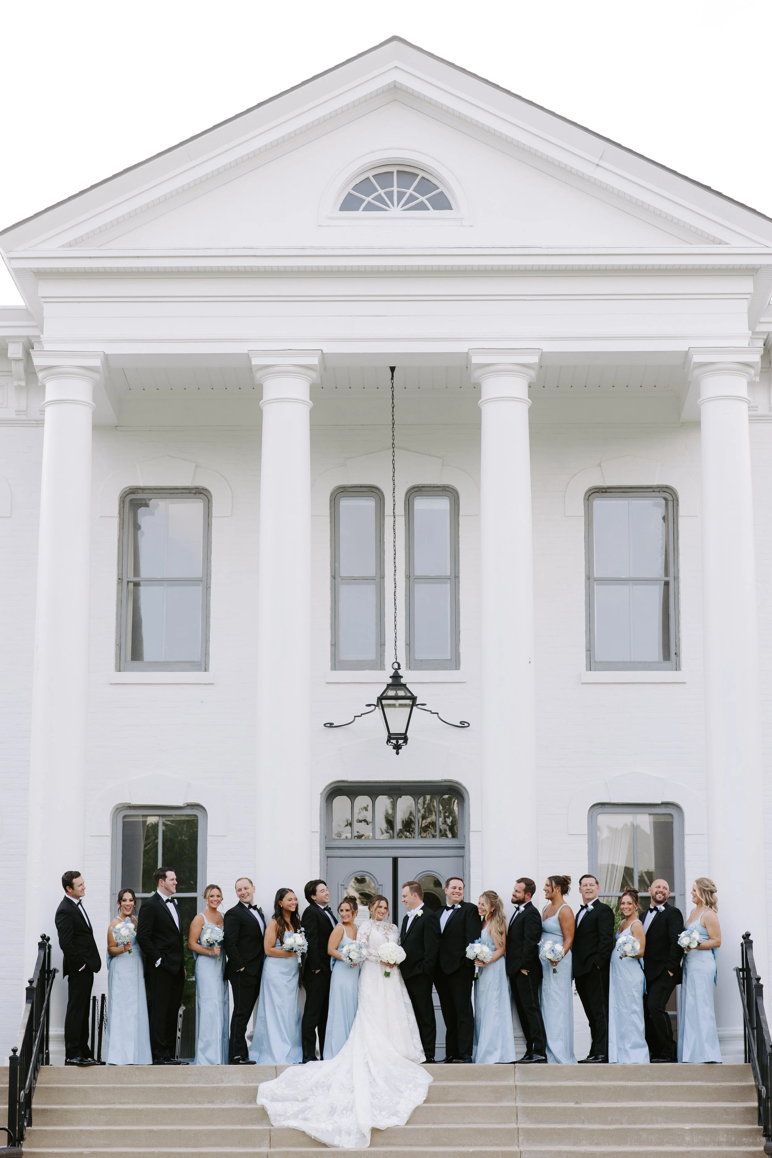 A wedding party standing on the steps of a white building with four large columns and an arch window at the top. The bride and groom are in the center surrounded by their bridesmaids and groomsmen dressed in formal attire.