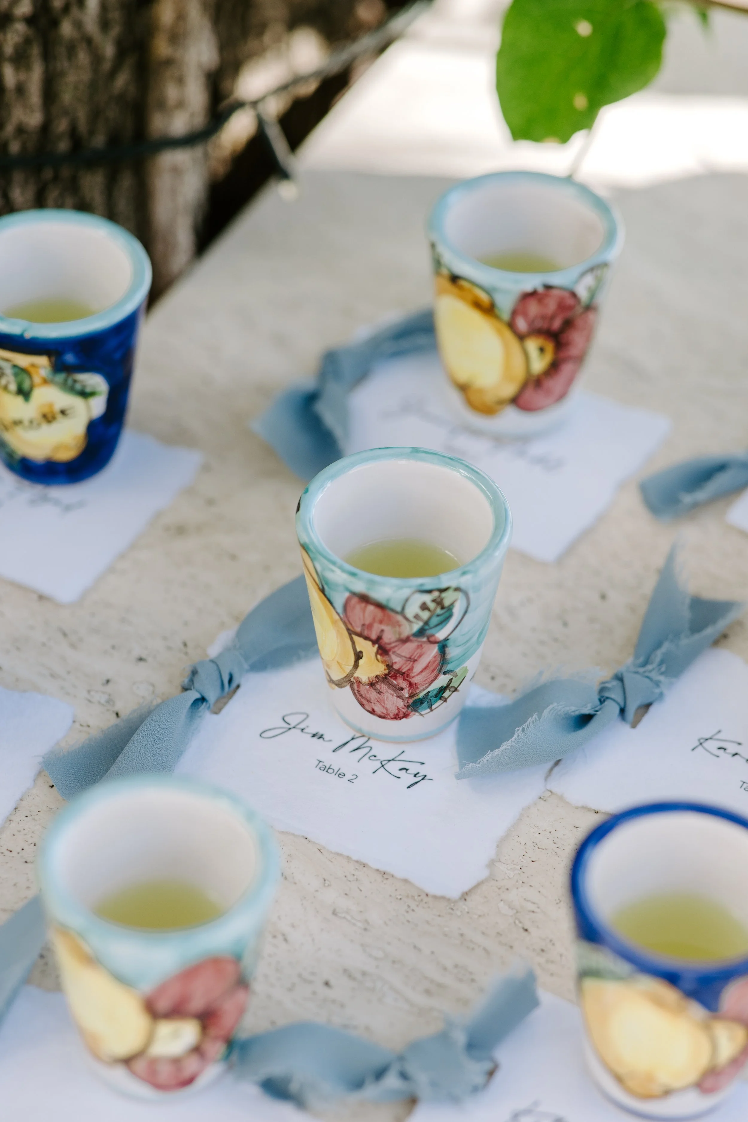 Colorful ceramic cups filled with a yellowish drink, arranged on a table with blue cloth decorations and handwritten name cards, outdoor setting with a blurred background.
