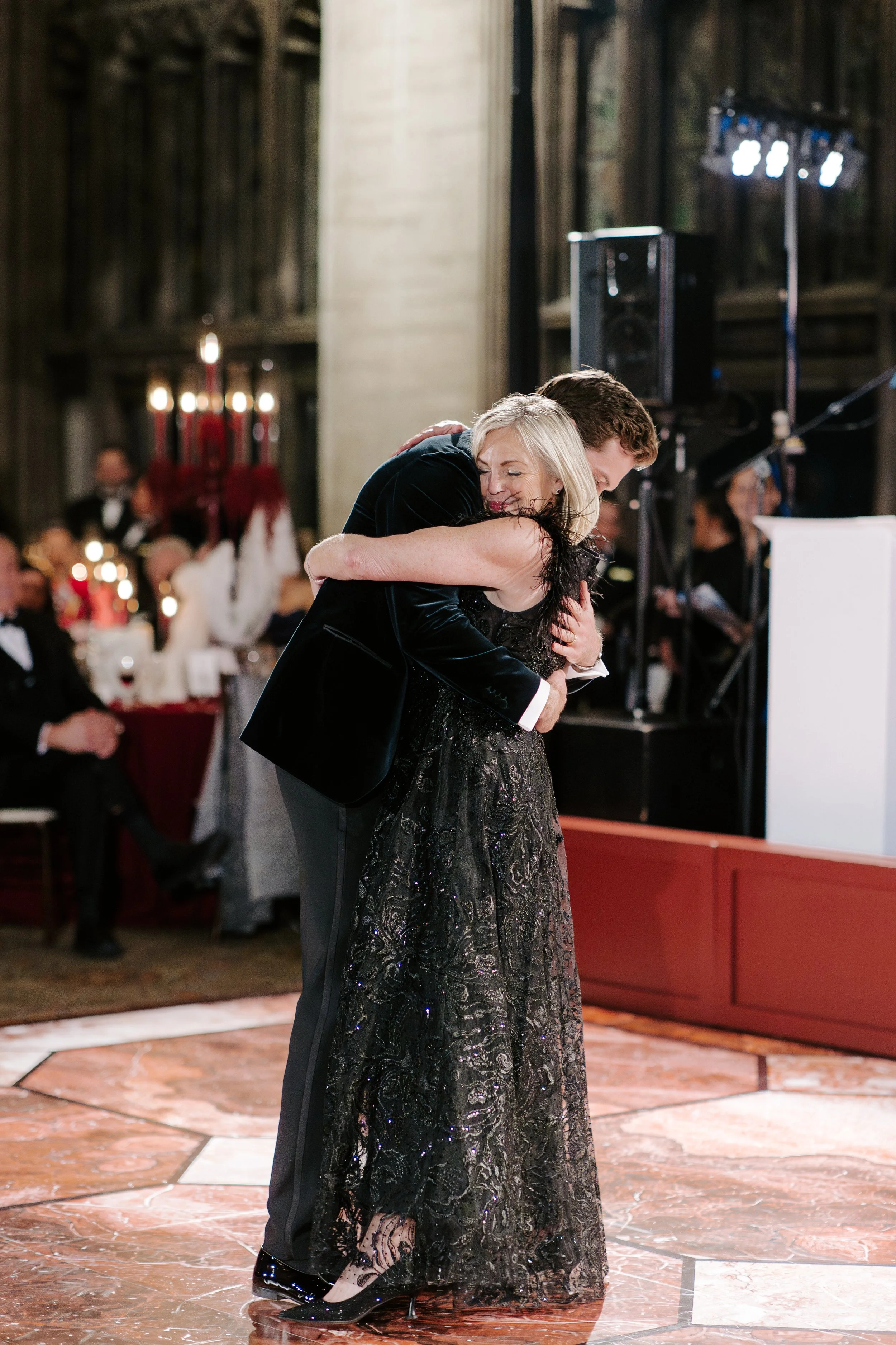 A man and woman hugging on a dance floor at a formal event, with people seated at tables in the background and stage lighting overhead.