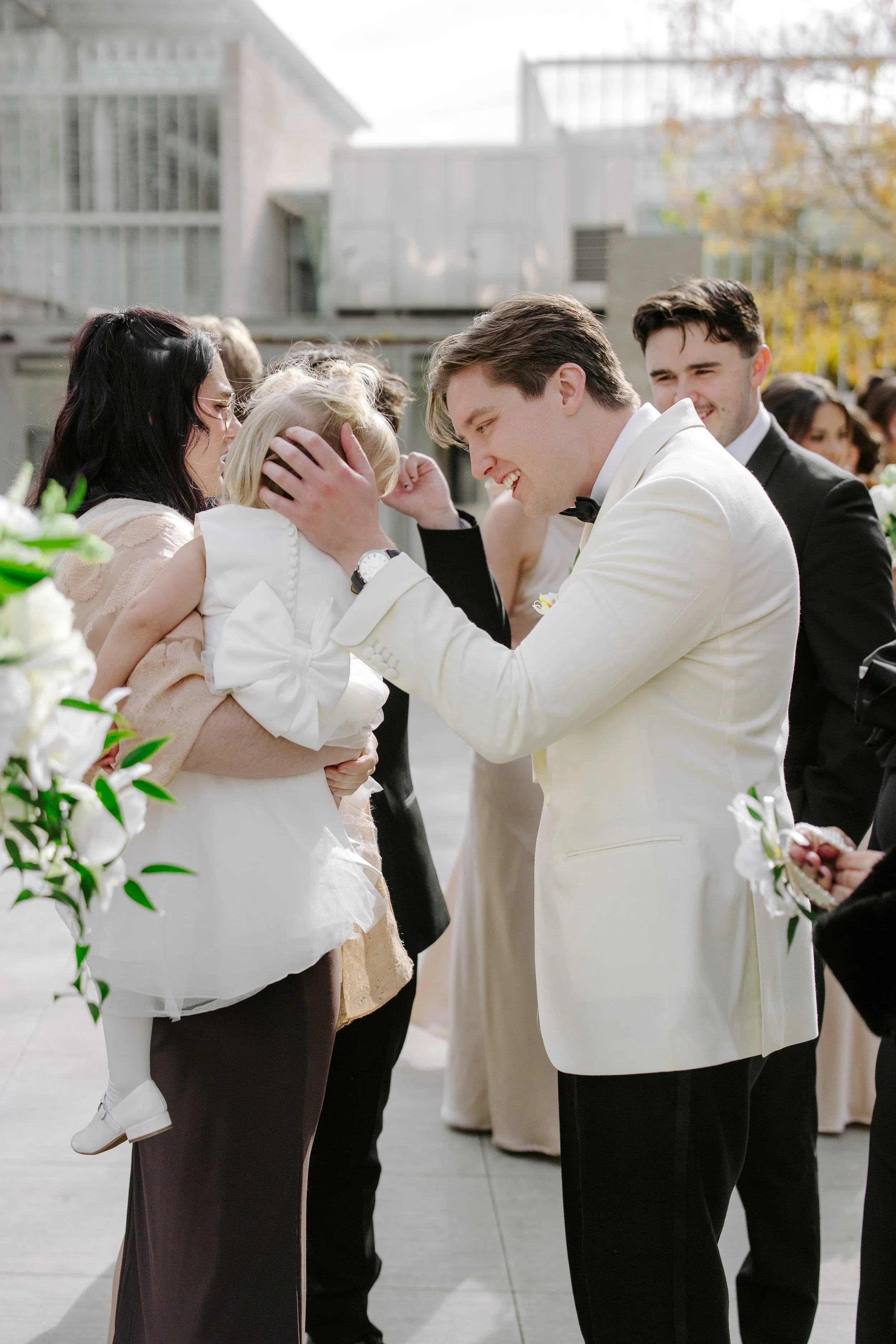 A wedding ceremony outdoors with a groom in a white tuxedo smiling and holding a young girl, possibly a flower girl, while a woman adjusts the girl's dress. Other people in formal attire are gathered around, with trees and a modern building in the ba