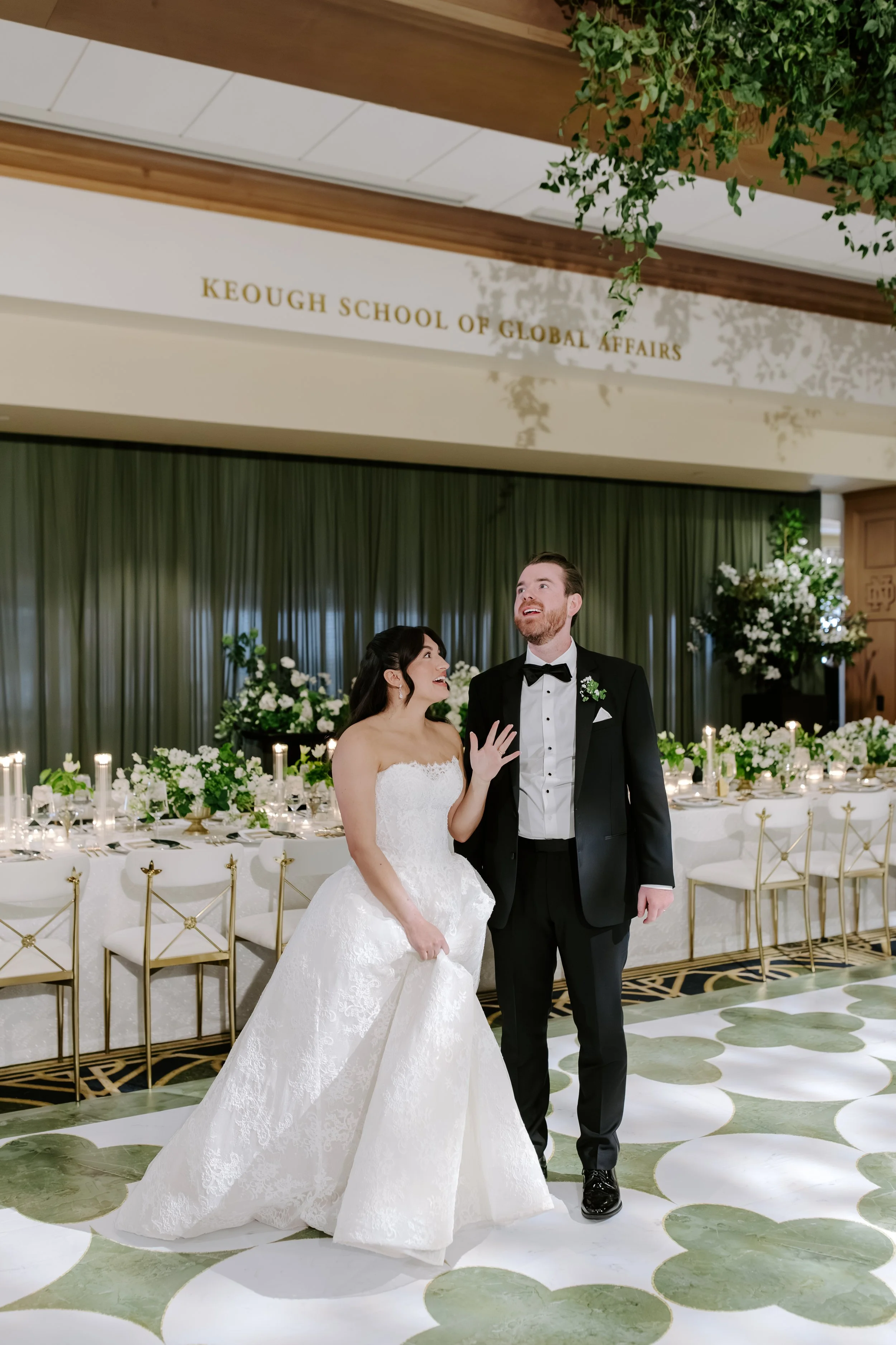 A bride and groom stand together in a decorated reception hall with green curtains and white floral arrangements. The bride wears a white strapless wedding gown, and the groom wears a black tuxedo with a bow tie. The bride is smiling and looking at t