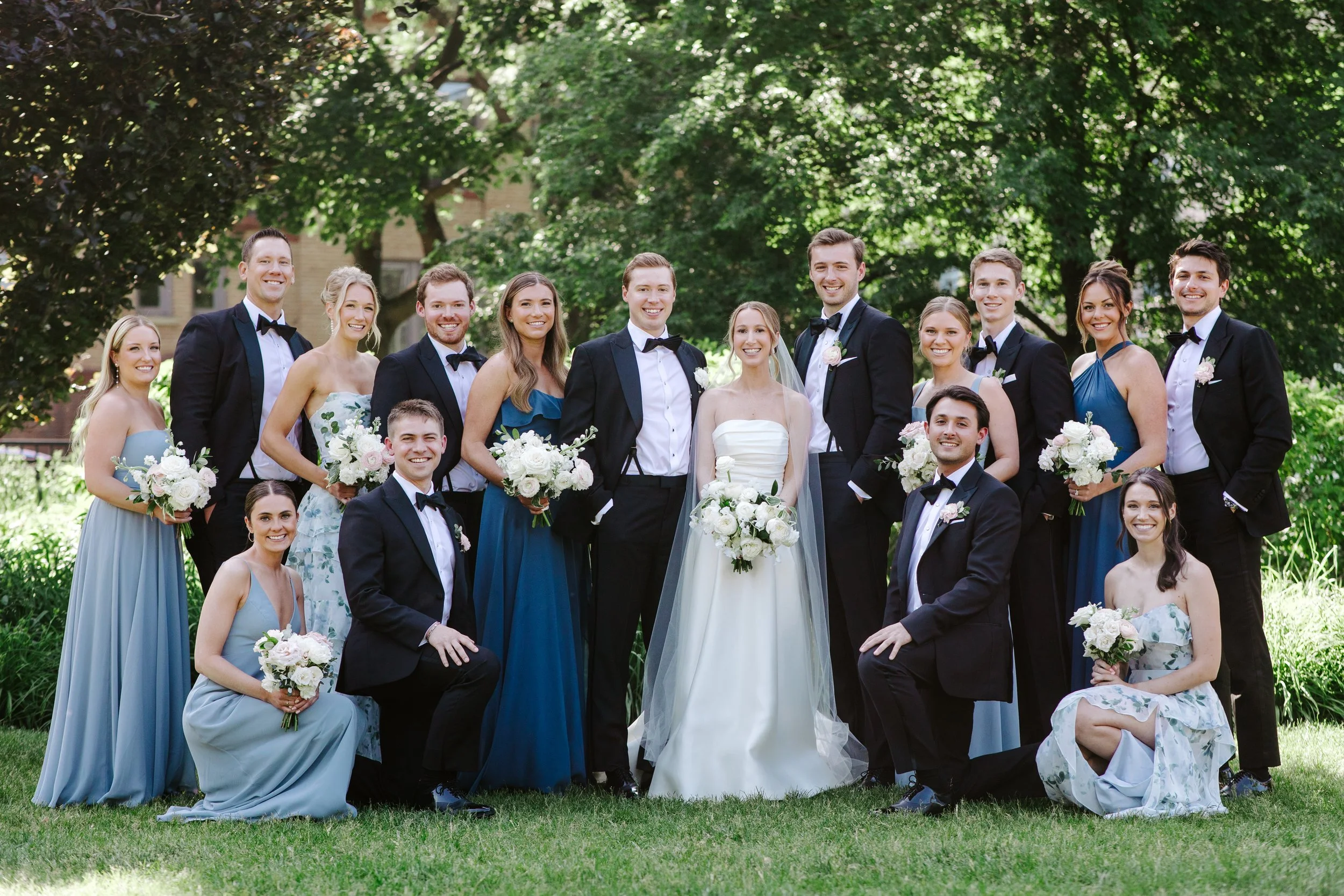 A large group of people, including men in tuxedos and women in floral and dressy outfits, gathered outdoors in a park-like setting with green trees in the background, celebrating at a wedding event.