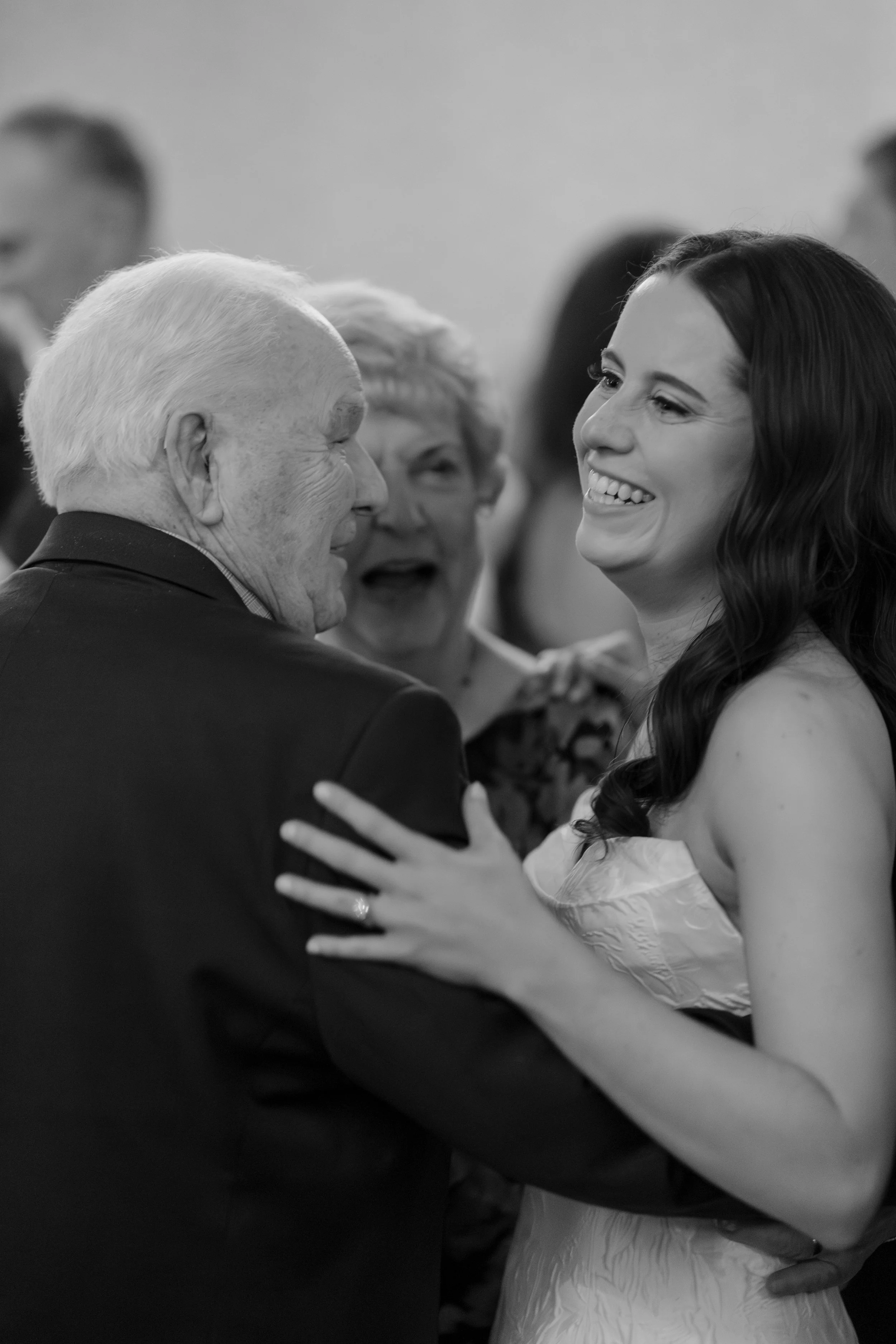 A black and white photograph capturing a joyful moment between a groom and bride during a wedding celebration. The groom, with white hair, is smiling and holding the bride close, while the bride, with long dark hair, is smiling broadly. They are surr