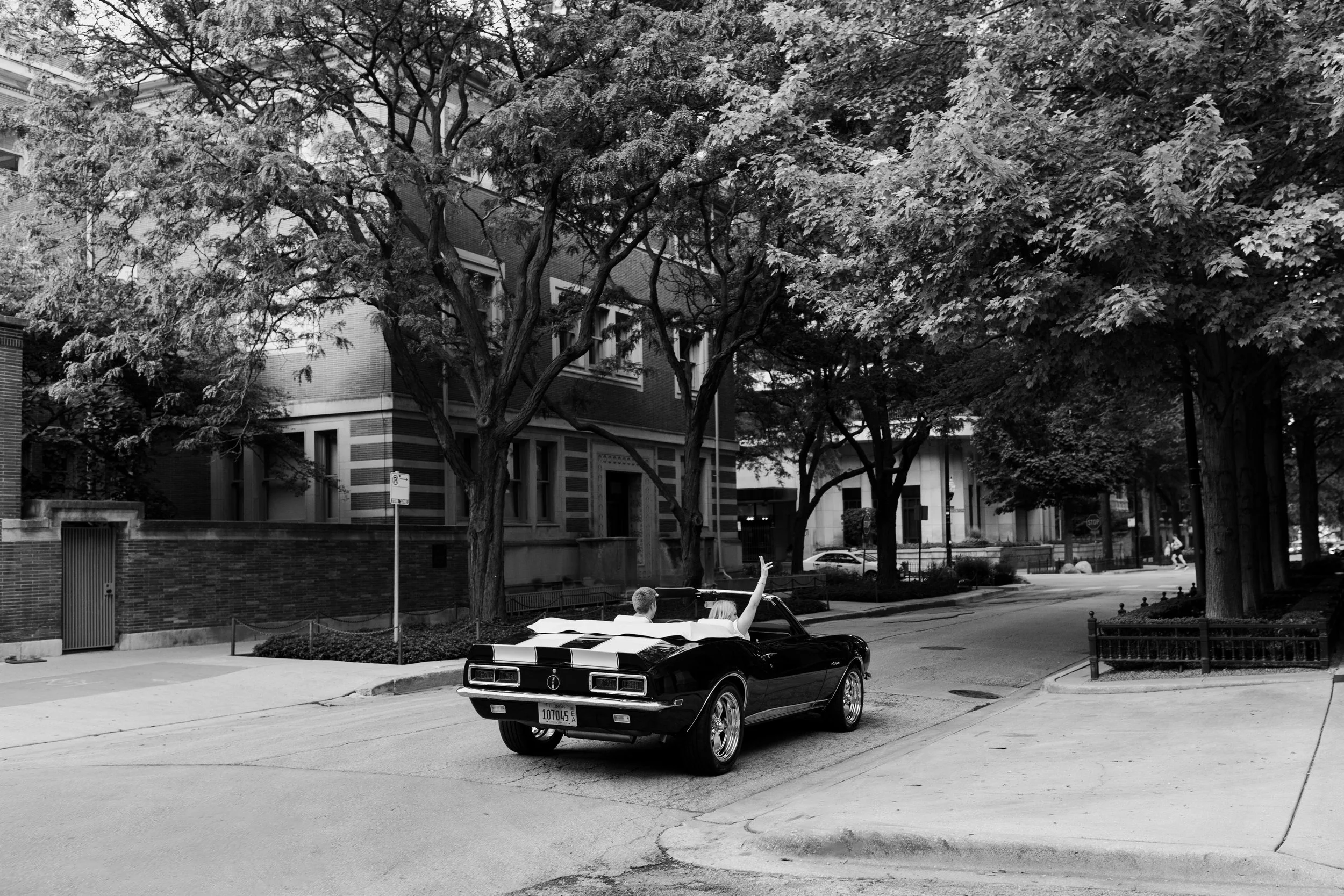 A black vintage convertible car with a man and woman inside, the woman raising her hand, driving down a tree-lined street in an urban neighborhood.