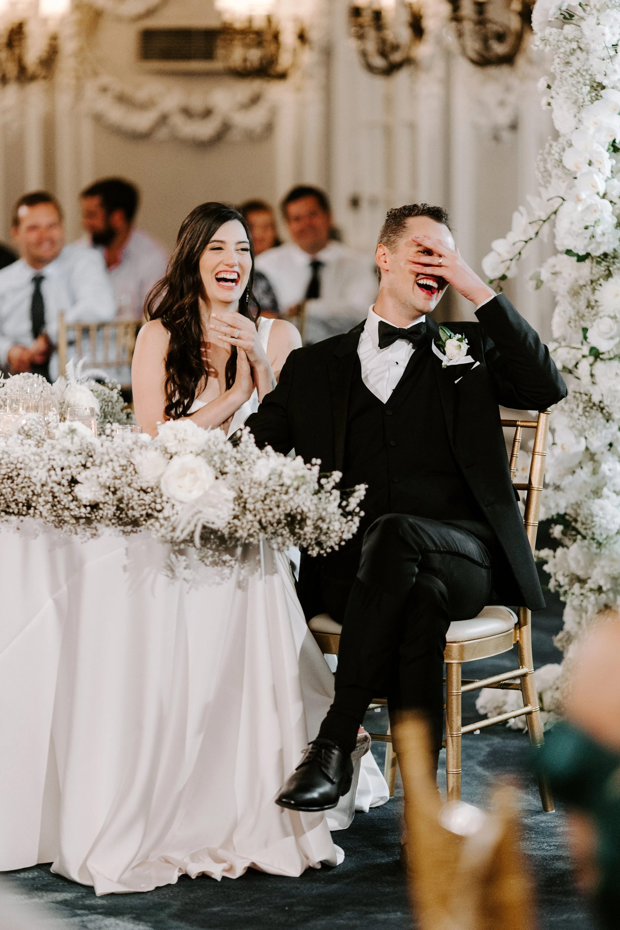 A bride and groom laughing during their wedding reception, sitting at a table decorated with white flowers.