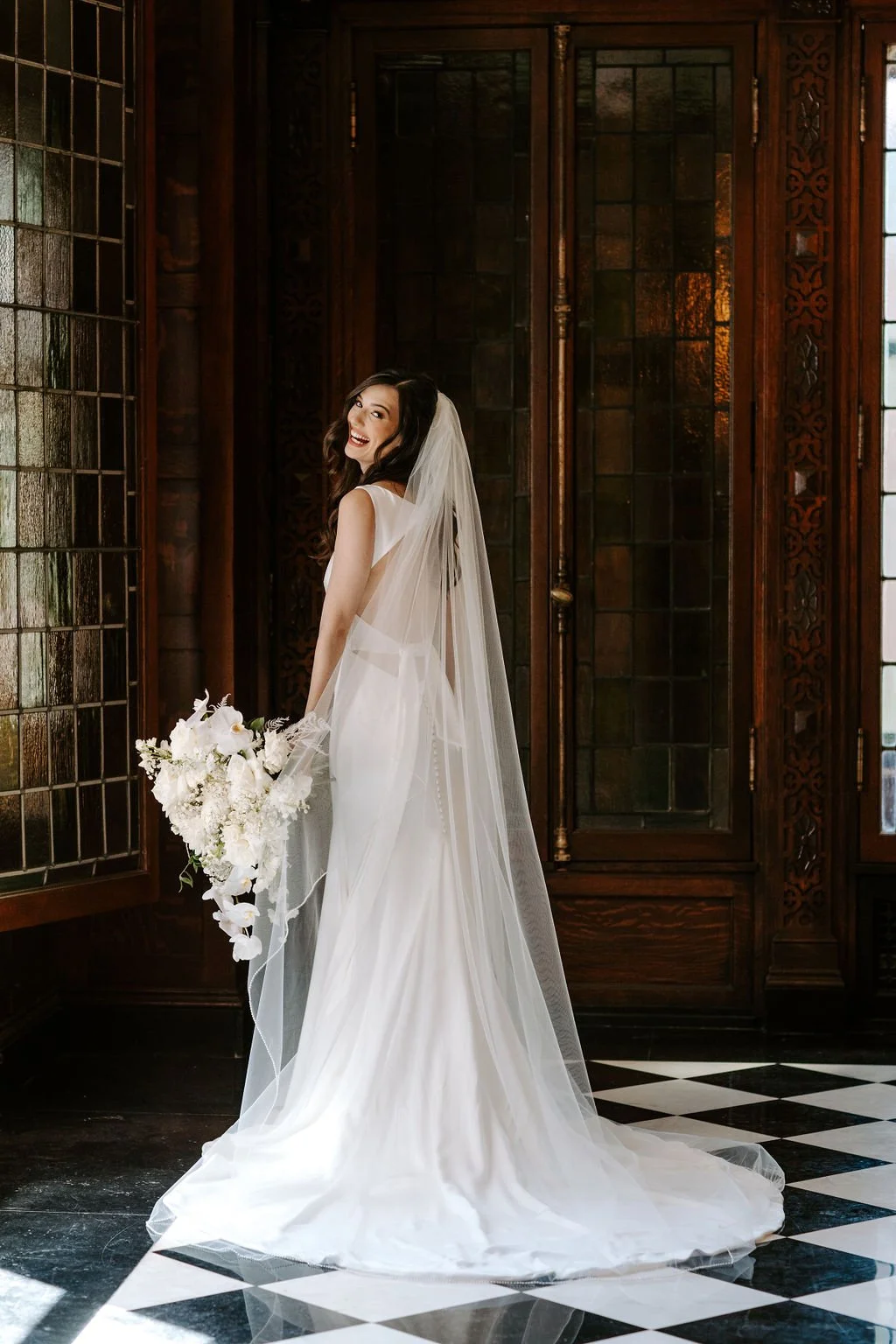 A bride in a white wedding dress holding a bouquet of white flowers, standing in a room with dark wood paneling and stained glass windows, smiling and turning her head.