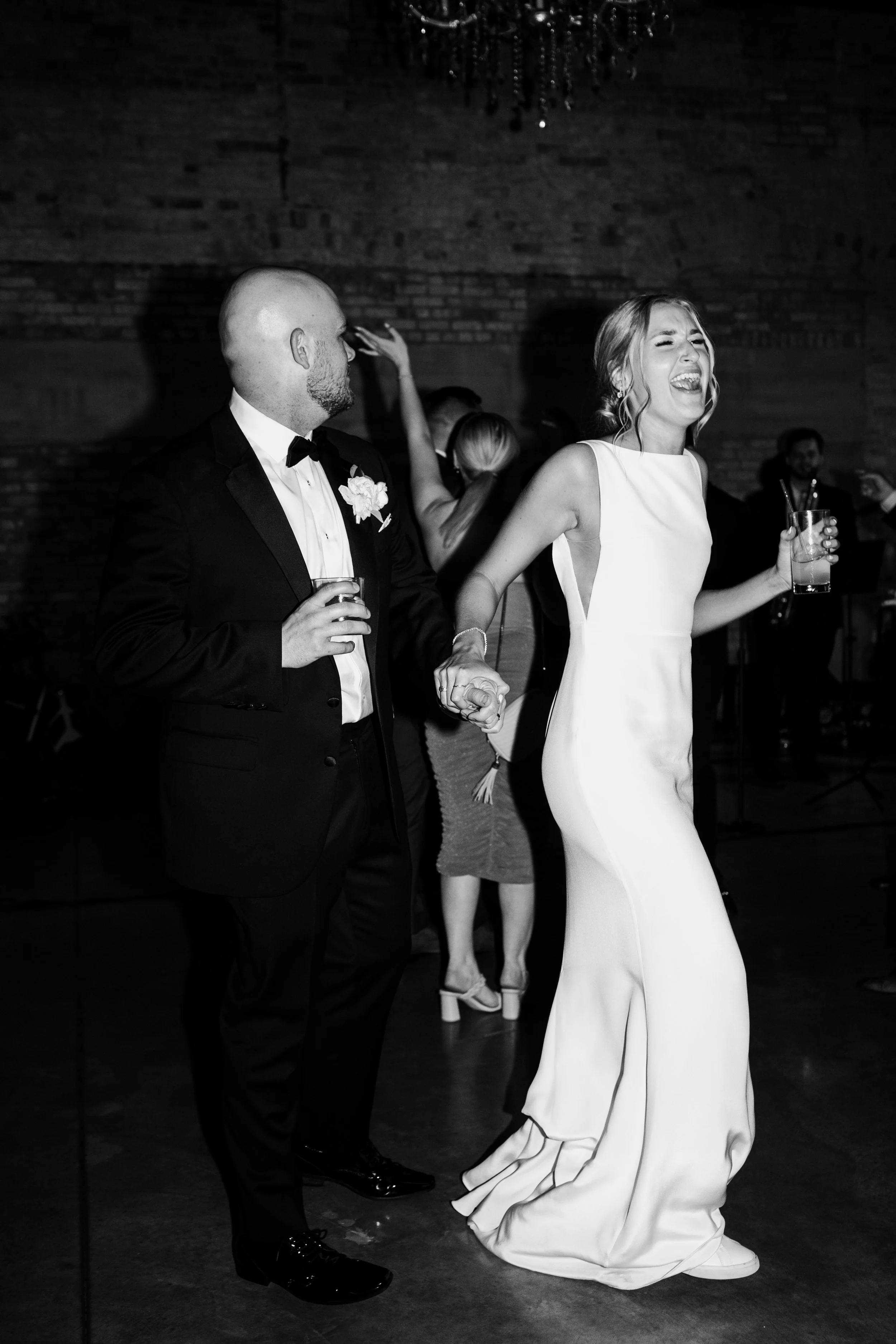 A bride and groom dancing at their wedding reception, holding hands and laughing, with other guests dancing in the background.