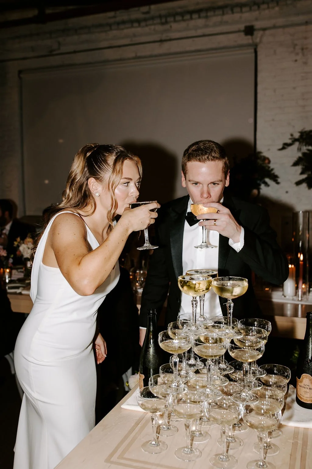 A woman and a man in formal attire are at a celebration, pouring and drinking champagne, with many glasses of champagne and bottles on a table in front of them.