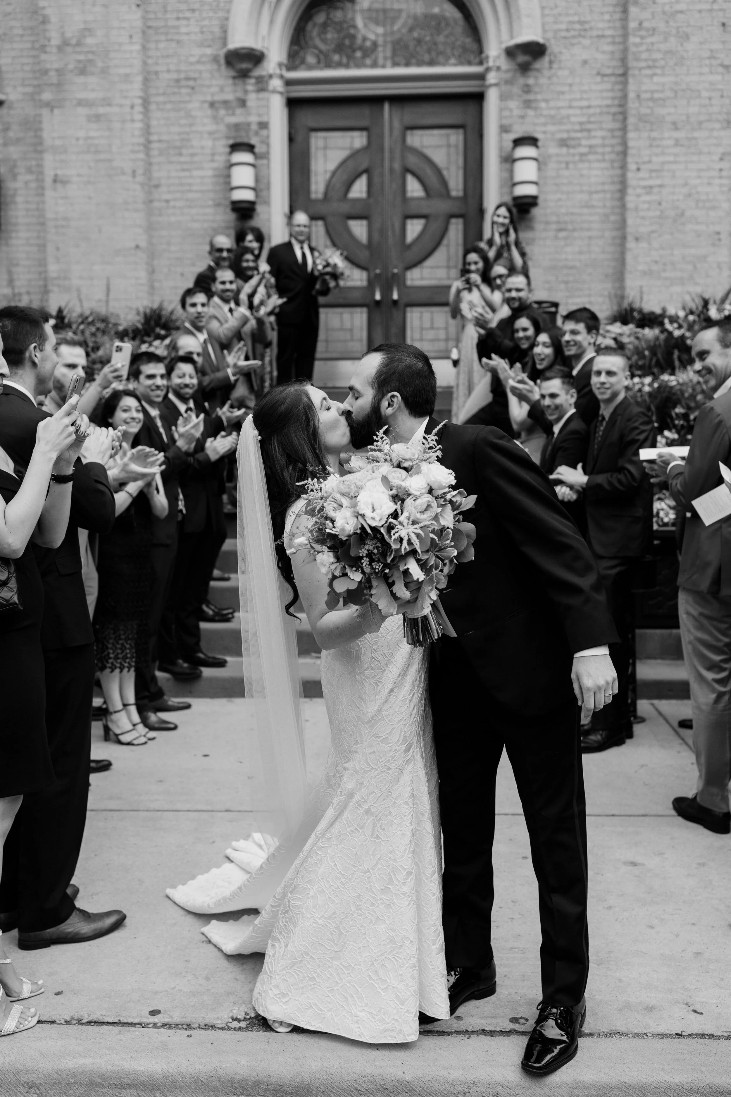A bride and groom are kissing outside a church after their wedding, surrounded by friends and family clapping and taking photos.