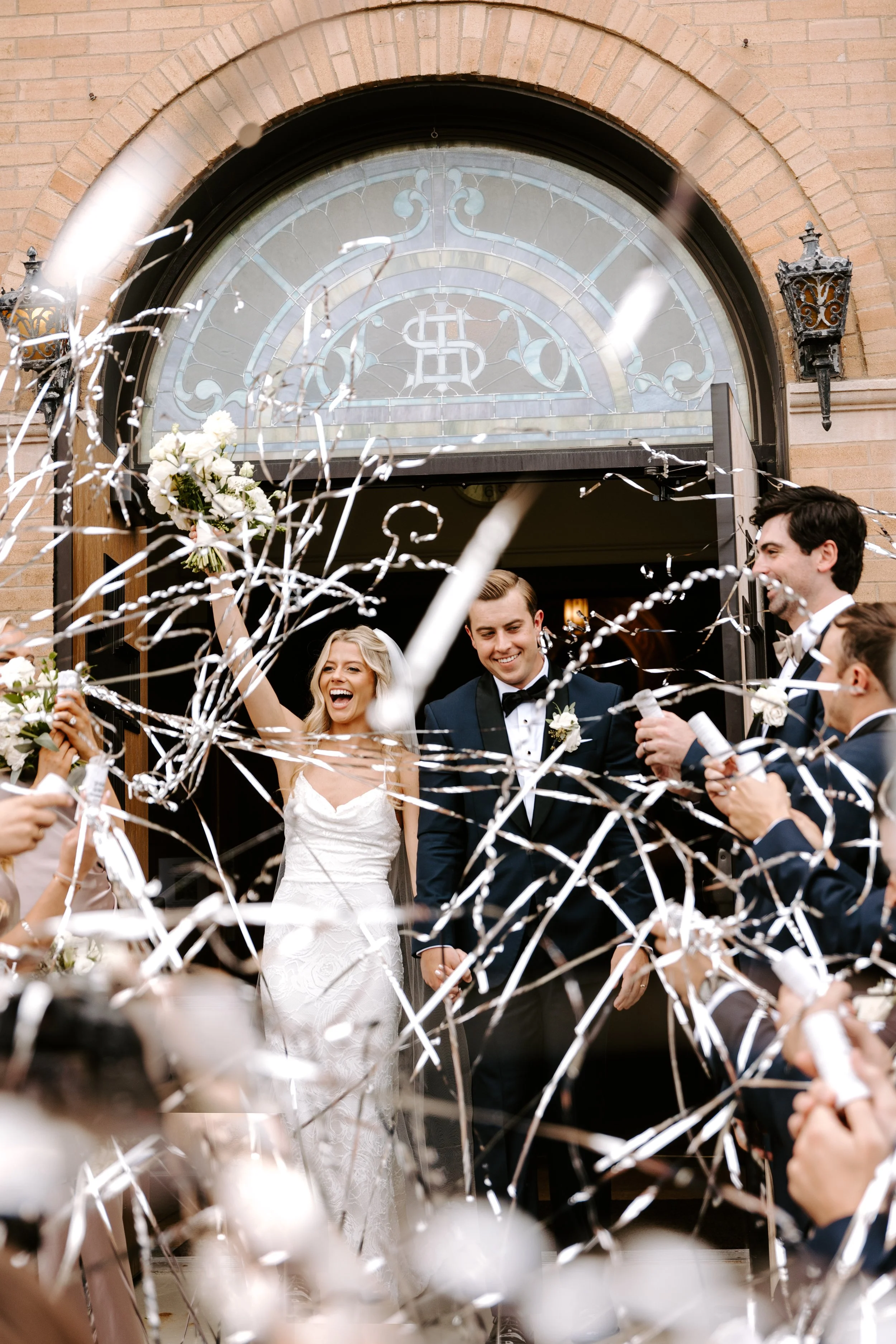 Bride and groom exiting a building on their wedding day, surrounded by guests throwing confetti and celebrating.
