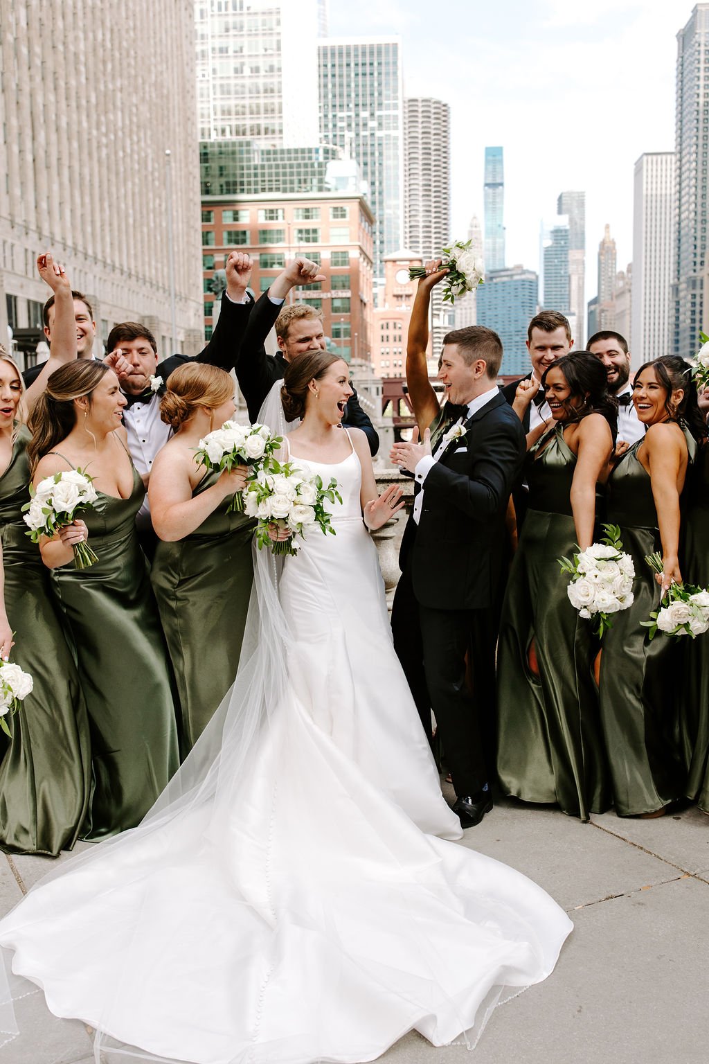 A wedding party celebrating outdoors in a city with tall skyscrapers, featuring a bride and groom in the center surrounded by bridesmaids and groomsmen holding flowers and cheering.