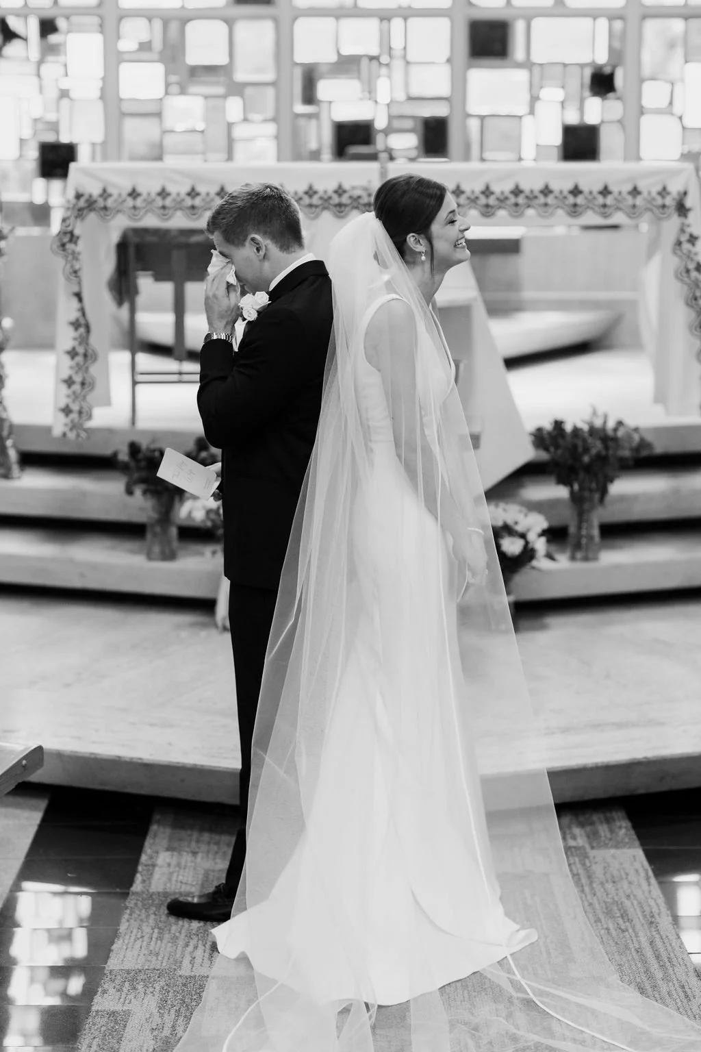 A bride and groom stand back-to-back during their wedding ceremony in a church, with the groom crying and the bride smiling.