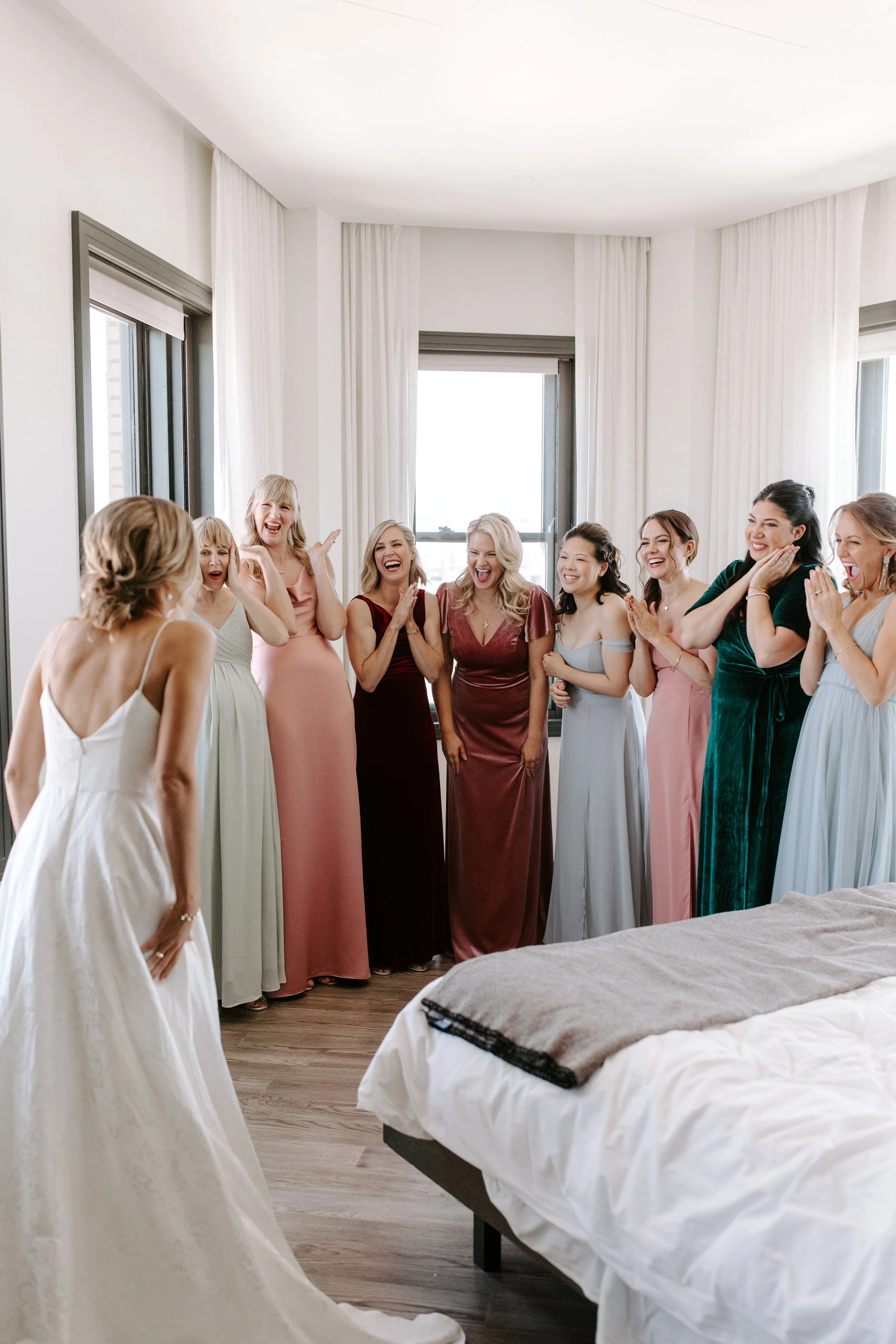 A group of women dressed in colorful gowns celebrating in a bright bedroom with large windows and white curtains.