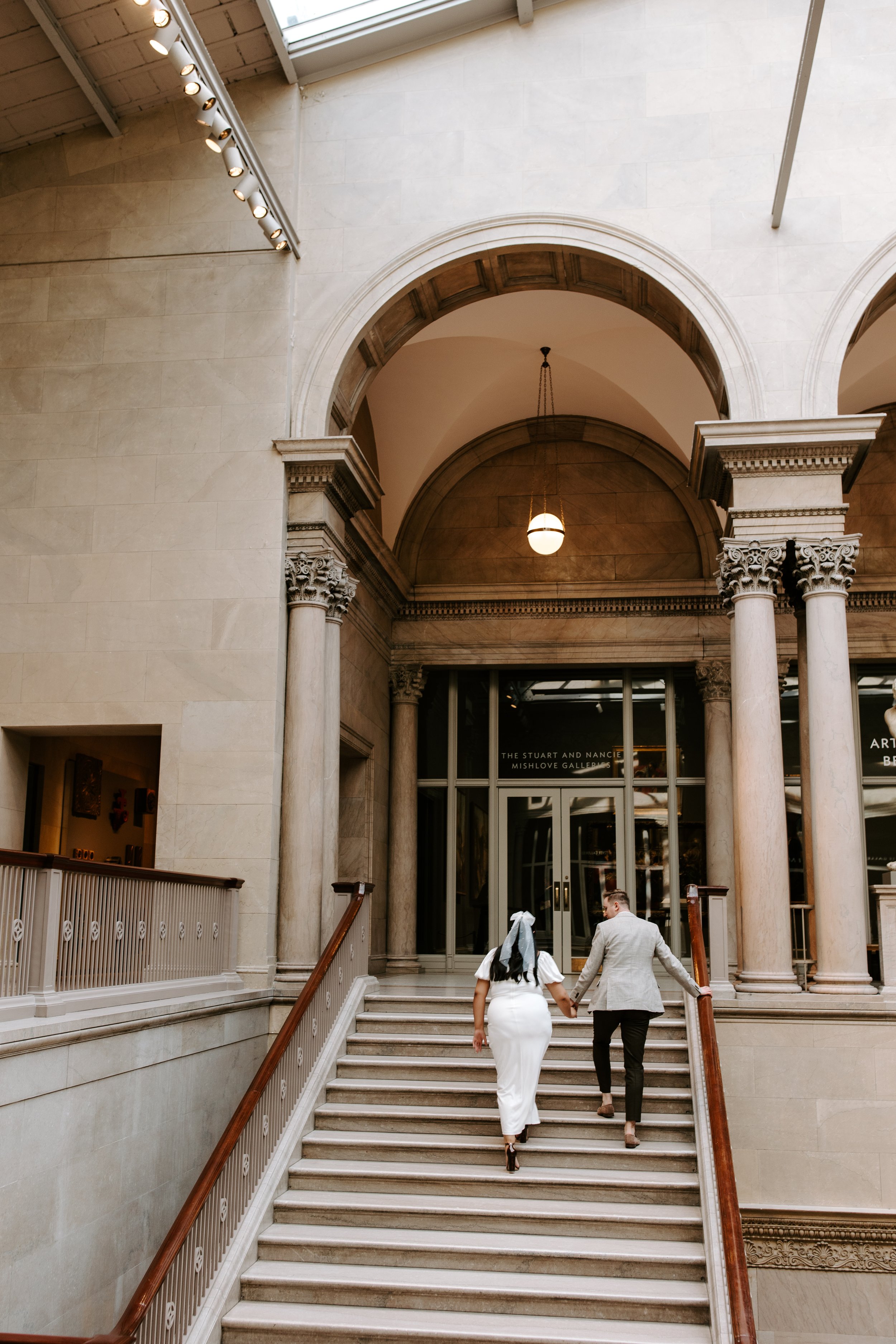 A man and woman holding hands and walking up a grand staircase inside an ornate building with classical architectural elements.