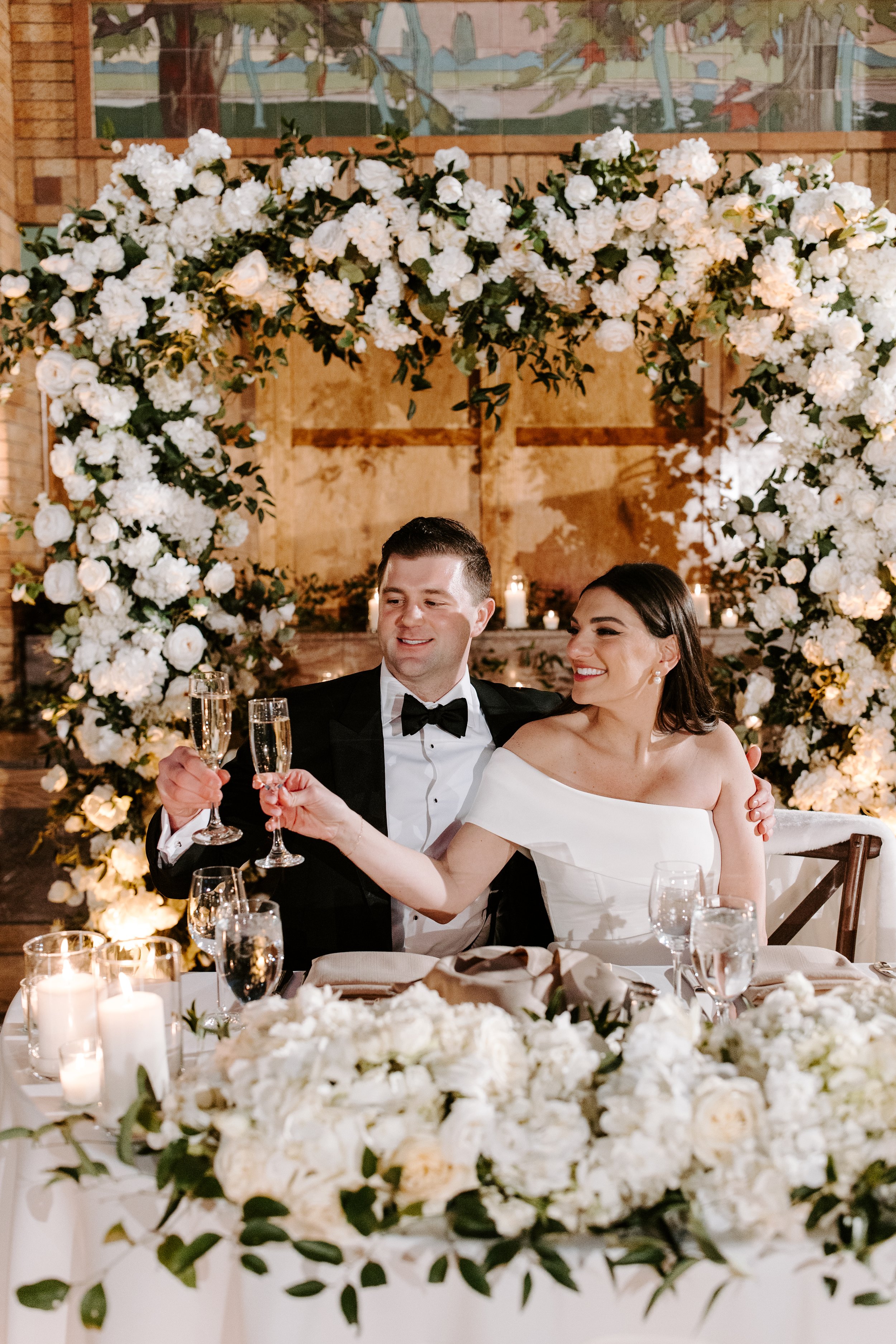 A newlywed couple celebrating at their wedding reception, sitting at a decorated table with lit candles and white floral arrangements, raising champagne glasses in a toast.