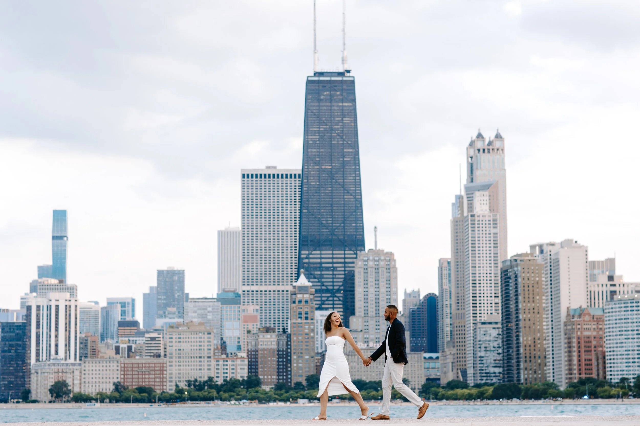A couple holding hands and walking along a city waterfront with the Chicago skyline, including the Willis Tower, in the background.