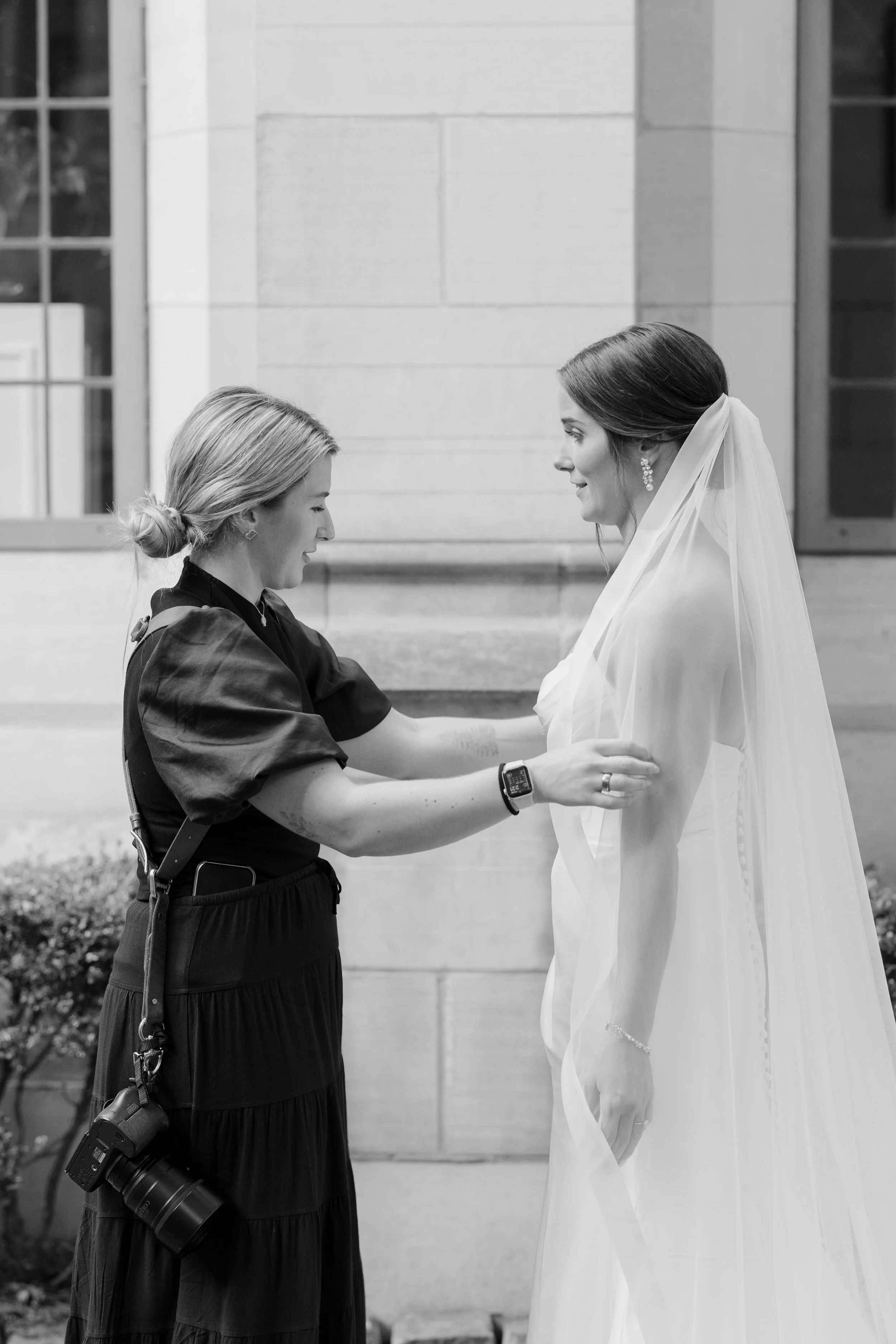 A woman in a wedding dress and veil stands outside in front of a light-colored building, smiling as another woman prepares her for her wedding, adjusting her dress or veil, with a camera hanging from her waist.