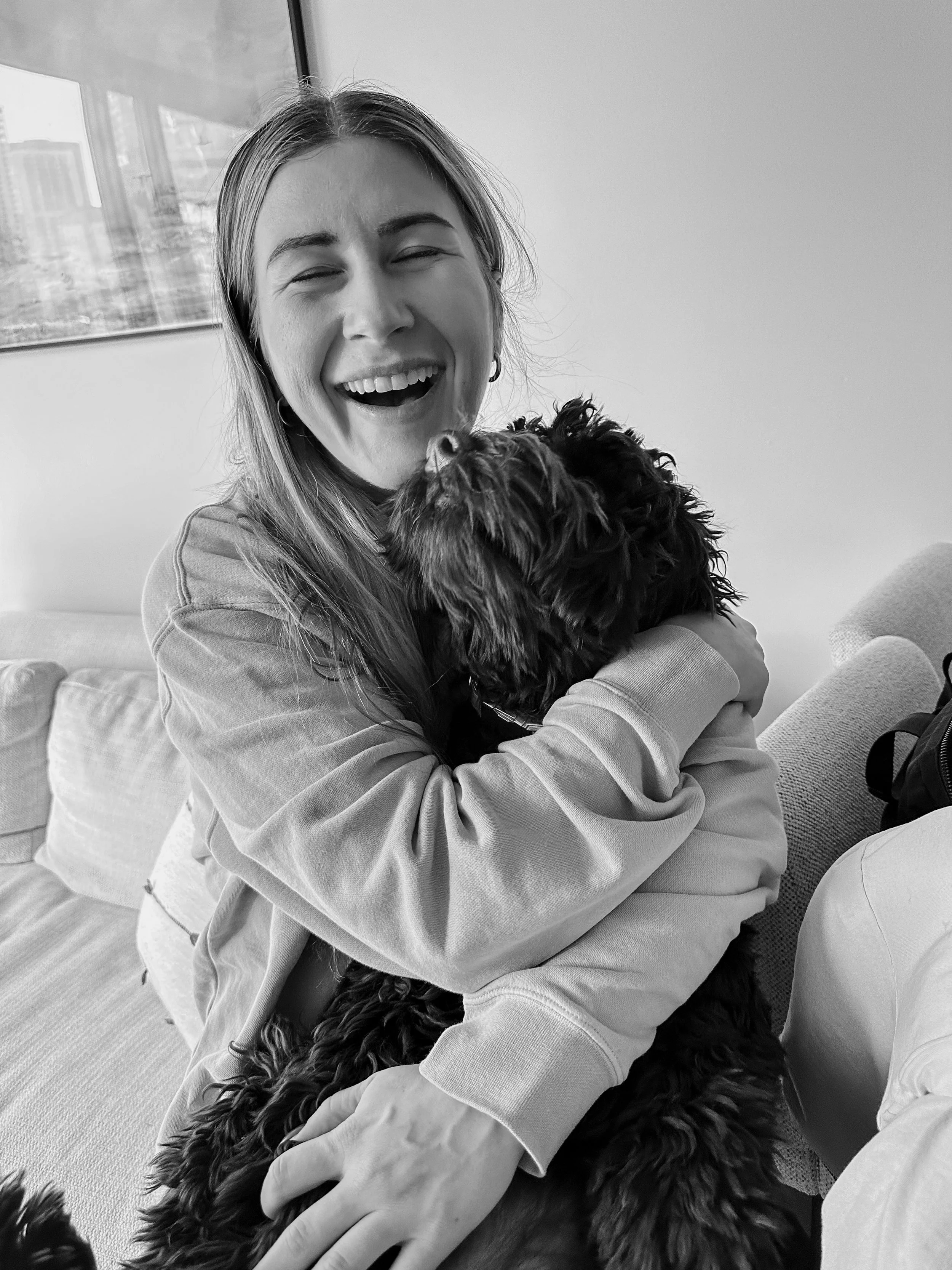 Black woman with long hair and hoop earrings joyfully hugging a black dog with curly fur in an indoor setting, smiling with eyes closed.