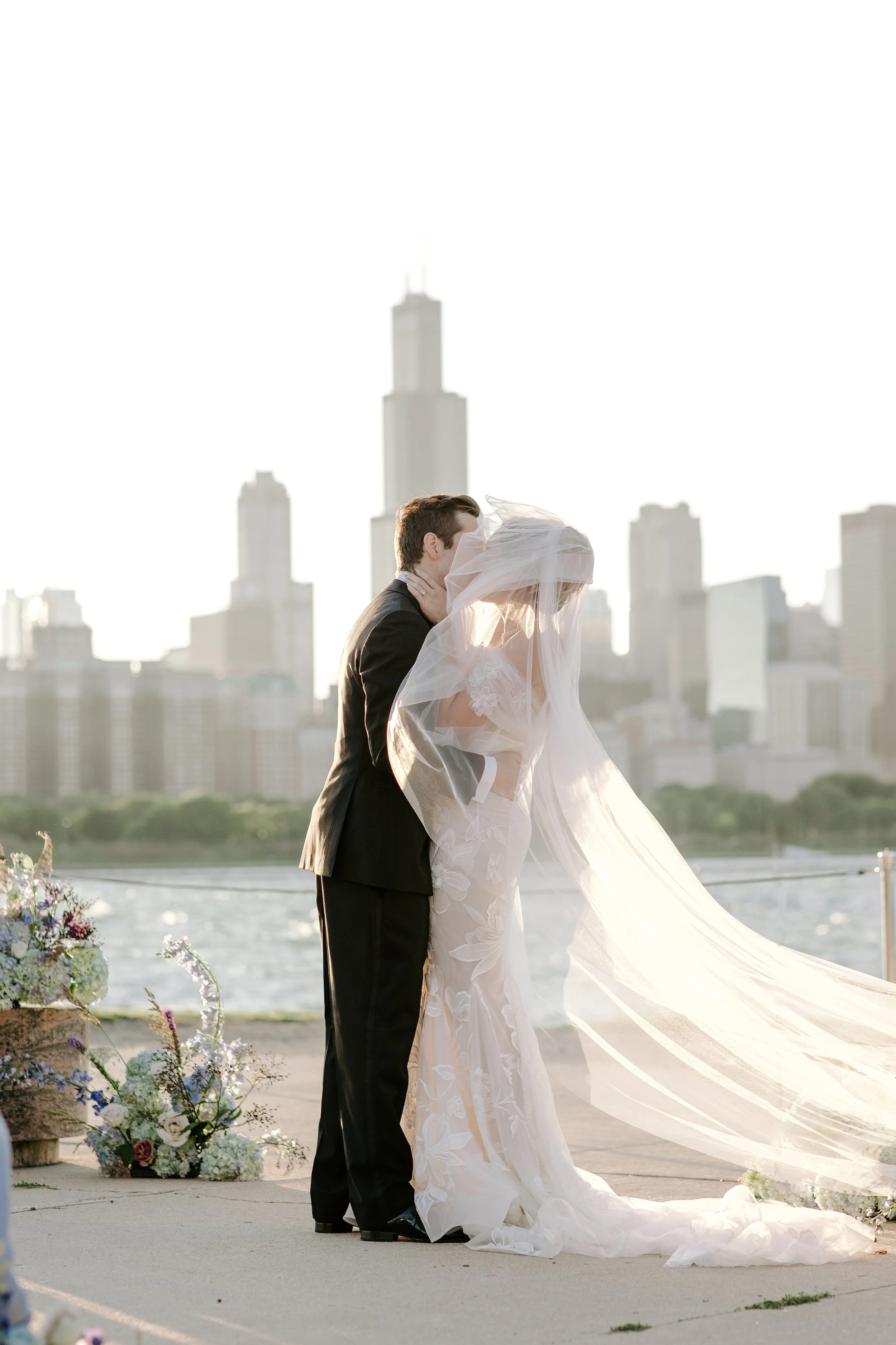 A bride and groom share a kiss during their outdoor wedding, with city skyline in the background and a river in the foreground.