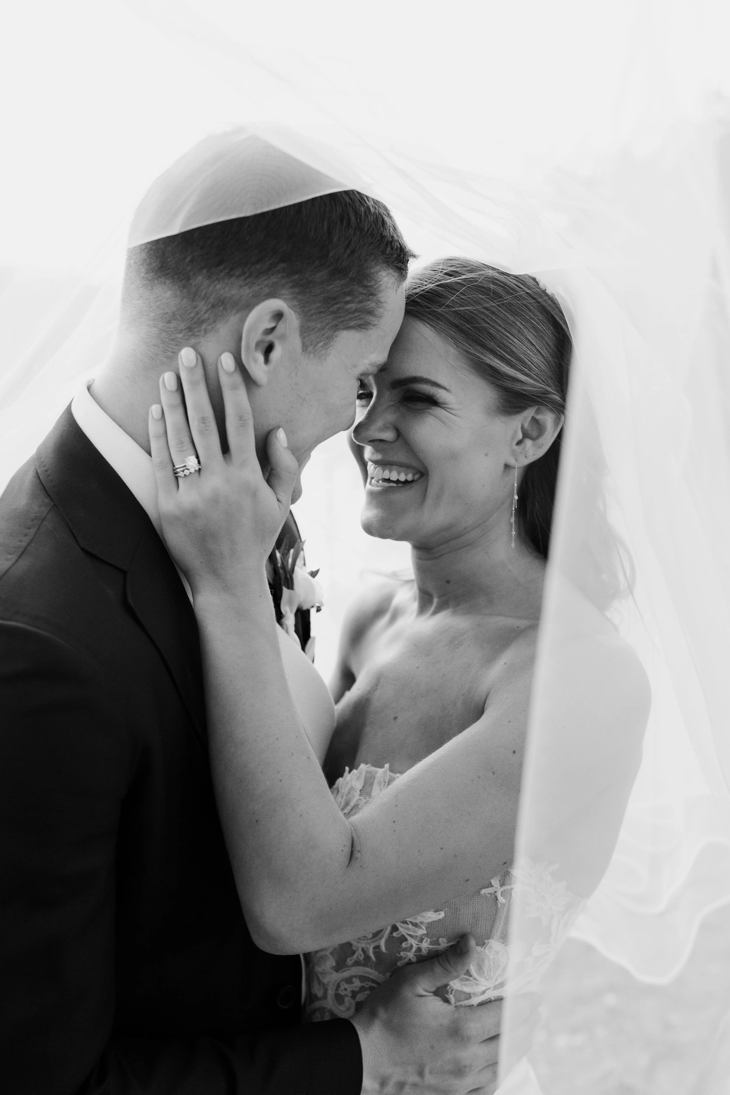 A black and white photo of a smiling bride and groom under a veil, with their faces close together, touching foreheads, during their wedding.