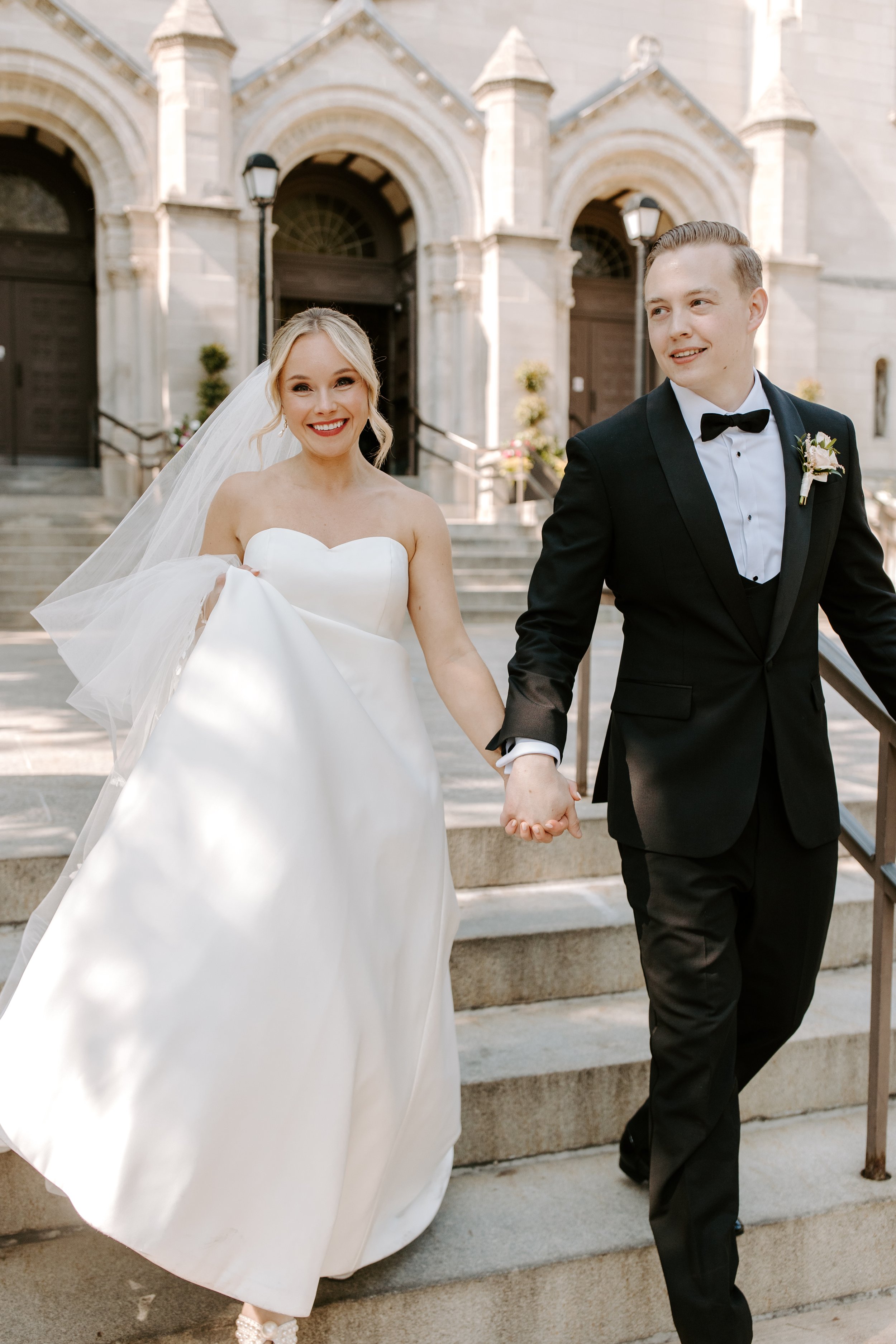 A bride and groom walking down steps outside a church after their wedding ceremony, holding hands and smiling.