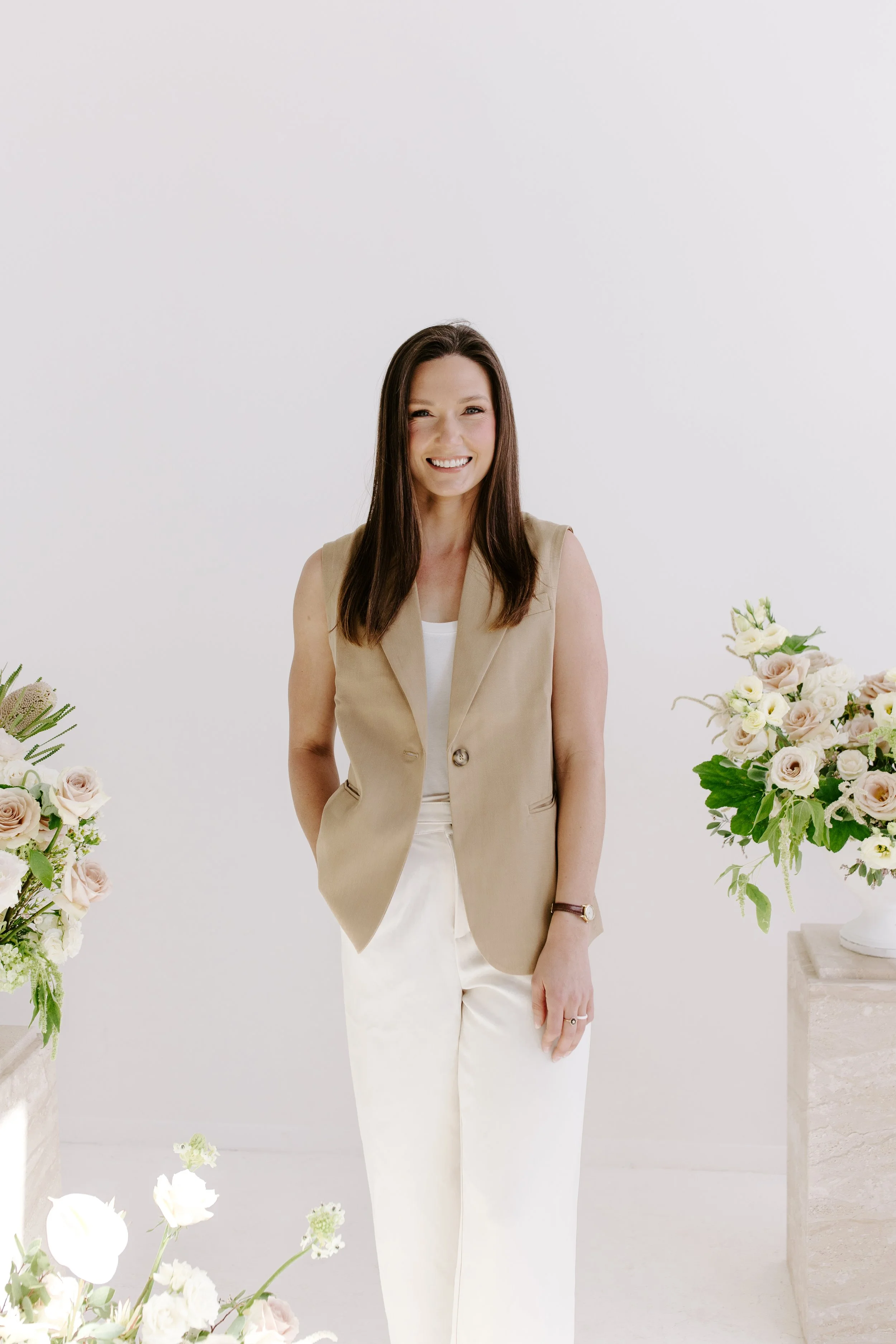 A woman smiling, standing in a white room with floral arrangements on either side.