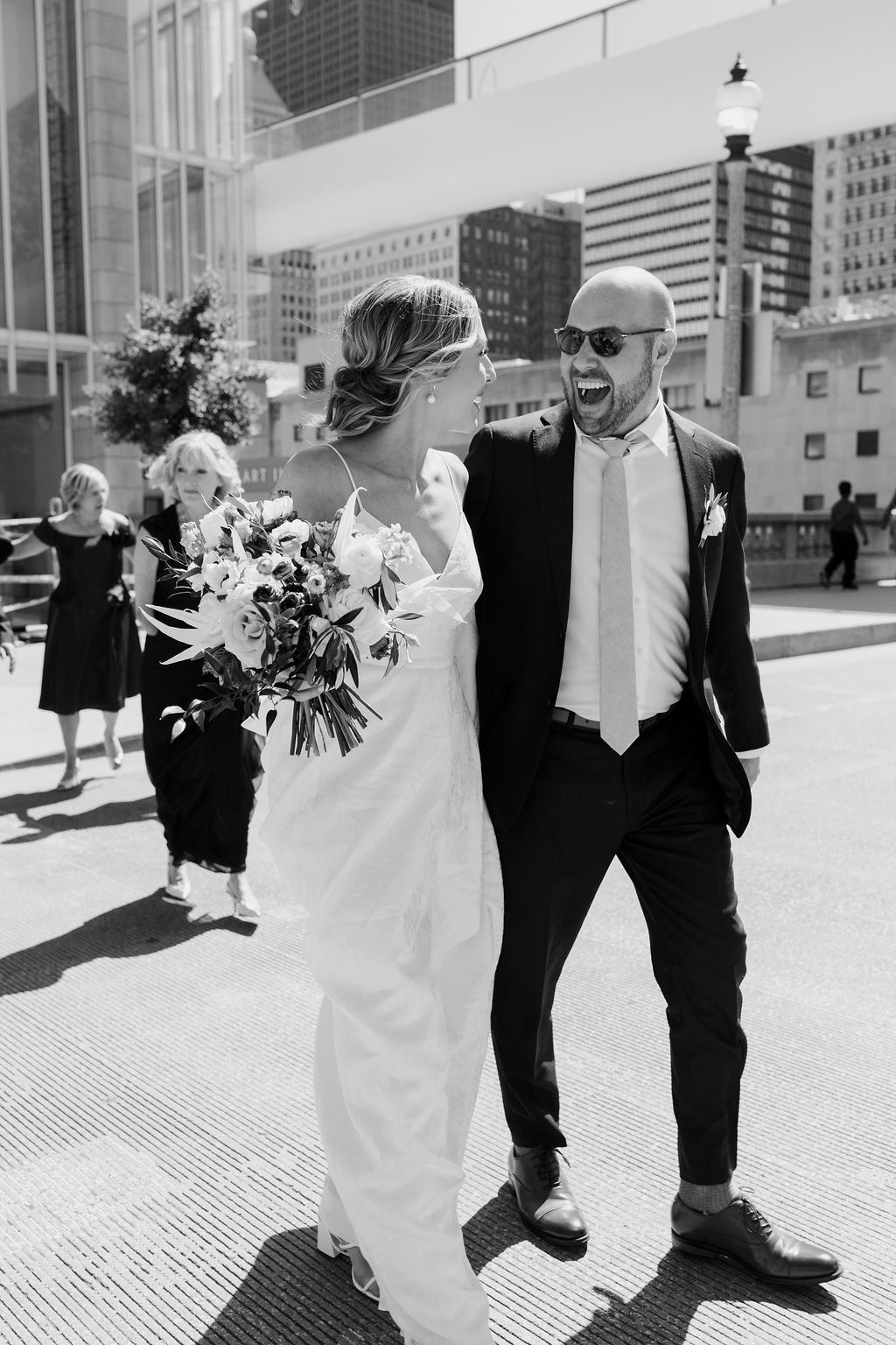 A black and white photo of a smiling wedding couple walking outdoors in an urban area, with tall buildings and a lamppost in the background. The bride is holding a bouquet, and the groom is wearing sunglasses.