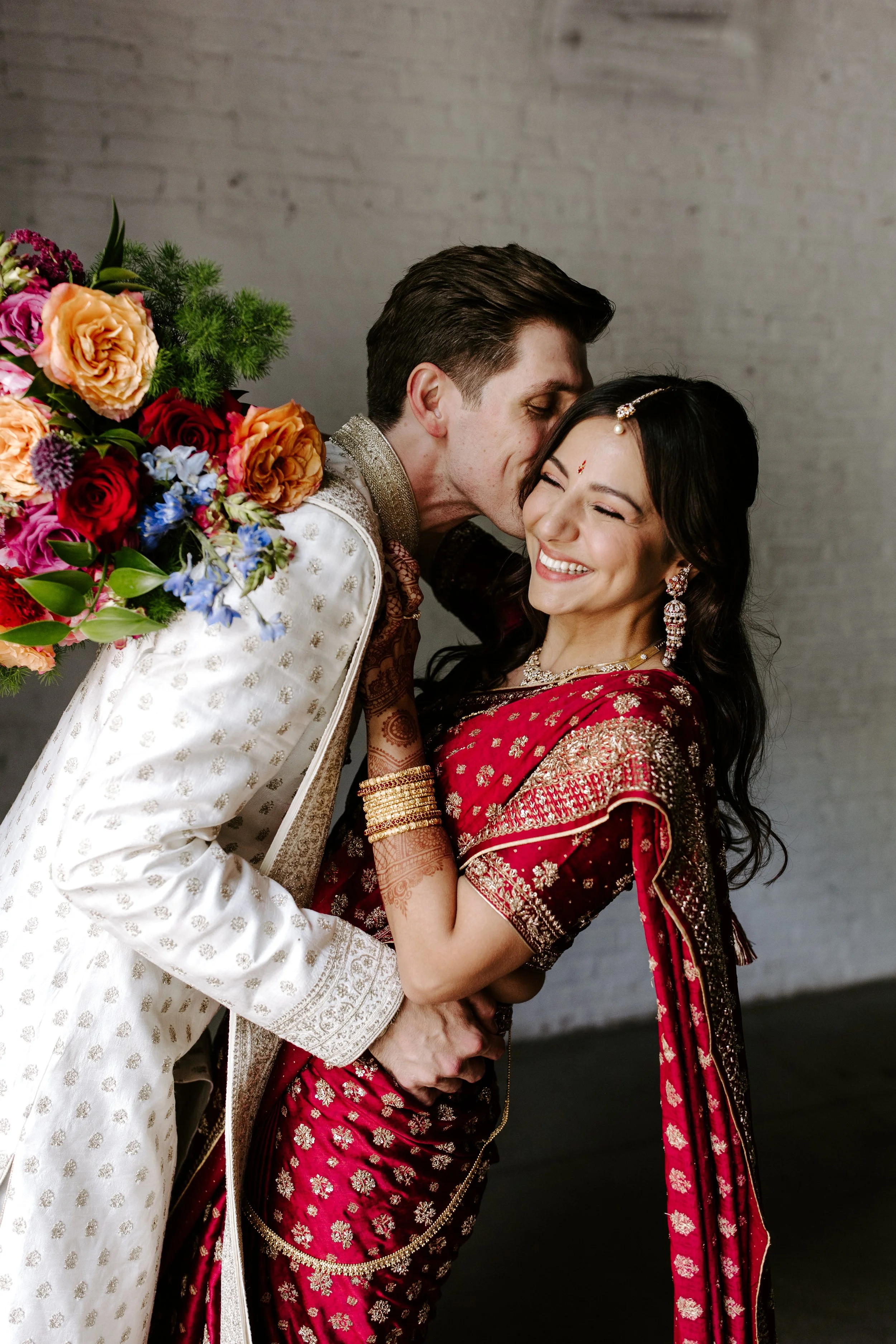 Bride and groom in traditional Indian attire sharing a joyful moment, with the groom kissing the bride on the forehead, holding hands, and smiling. The bride is dressed in a red saree with gold embroidery, adorned with jewelry, and henna on her hand.