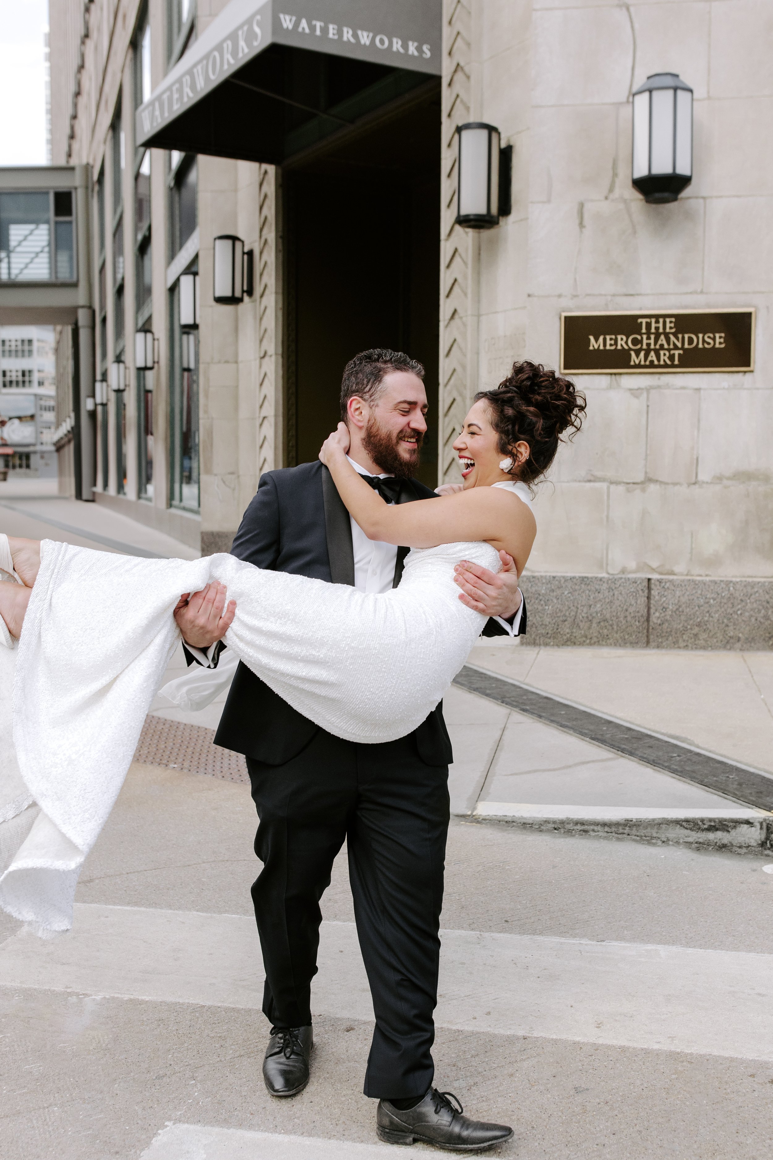 A happy couple in wedding attire, with the man in a black tuxedo and the woman in a white wedding dress, smiling as he carries her outside a building with a sign that reads 'The Merchandise Mart' in an urban setting.