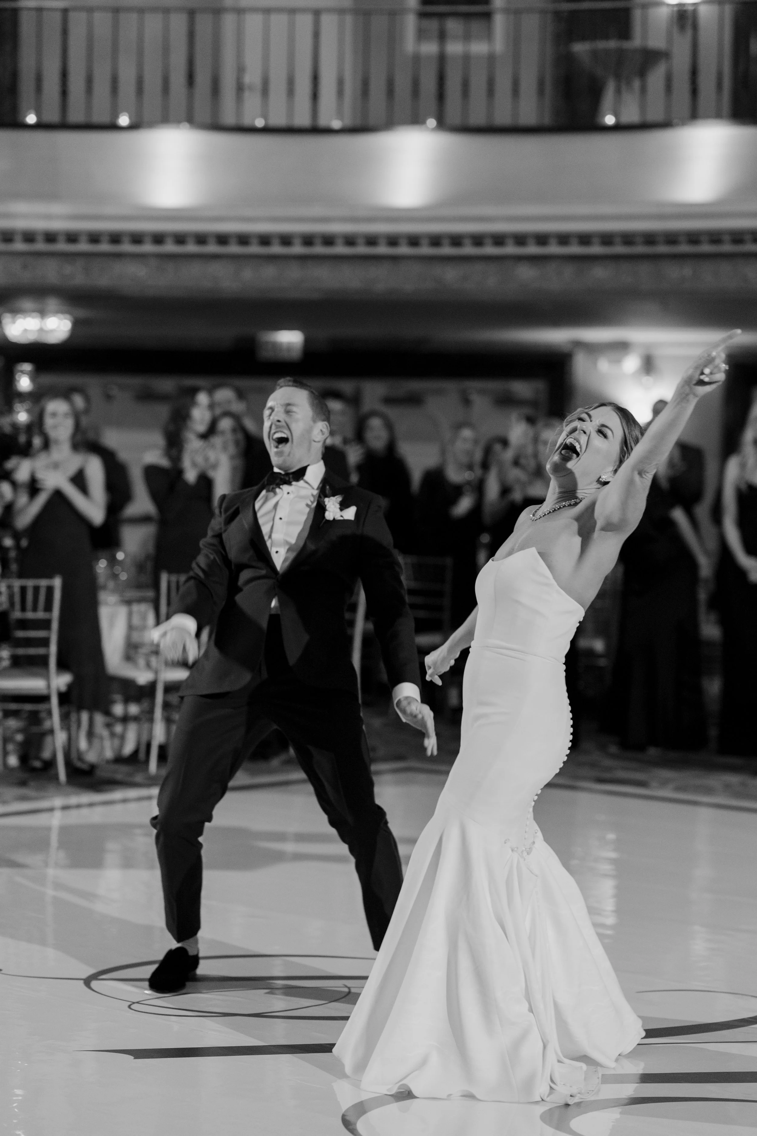 A bride and groom dancing joyfully at their wedding reception, with guests watching and smiling in the background.