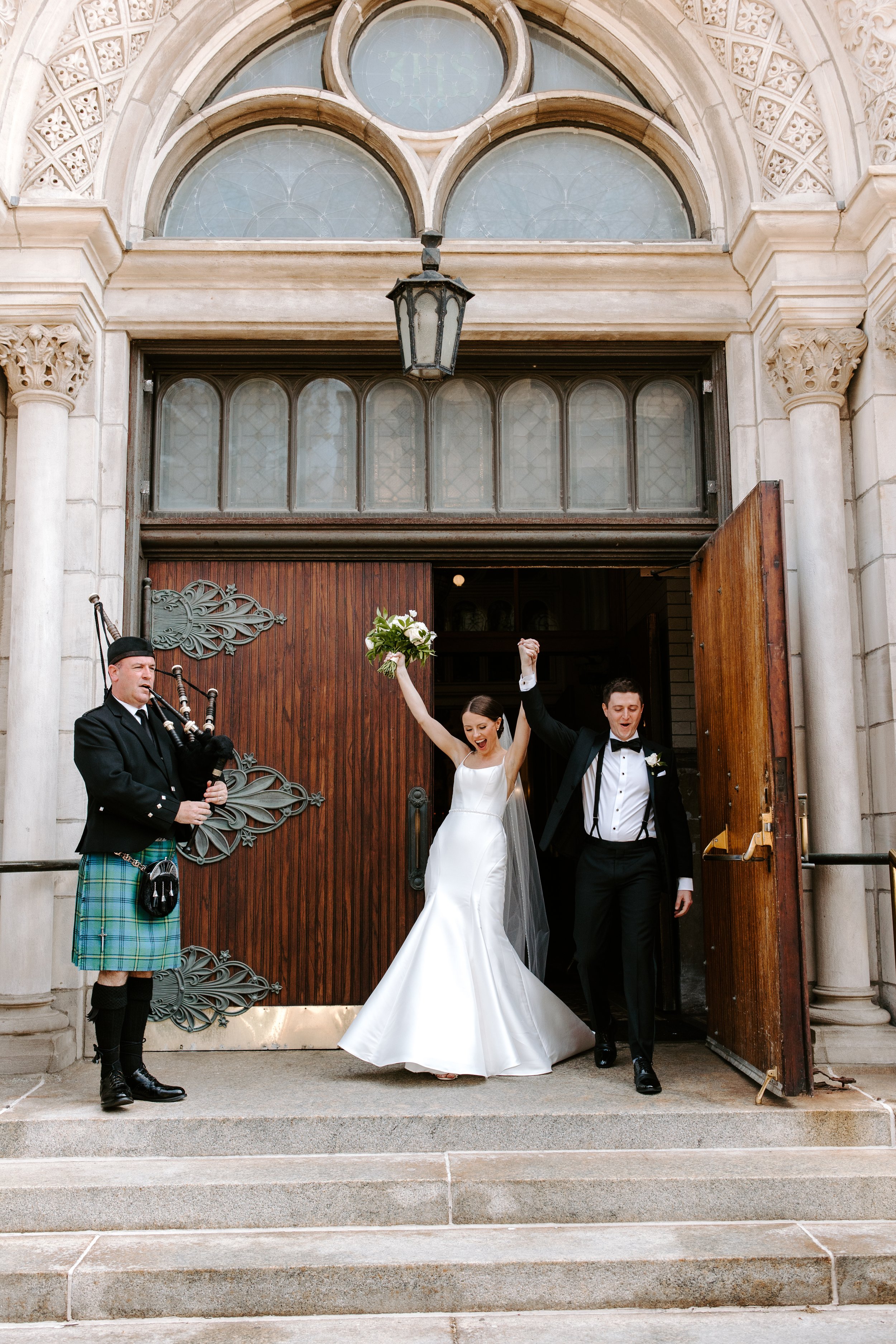 A newlywed couple exiting a church, celebrating with raised arms. The bride is in a white wedding gown holding a bouquet, and the groom is in a black tuxedo. A pipe band, dressed in traditional Scottish attire, stands to the side.