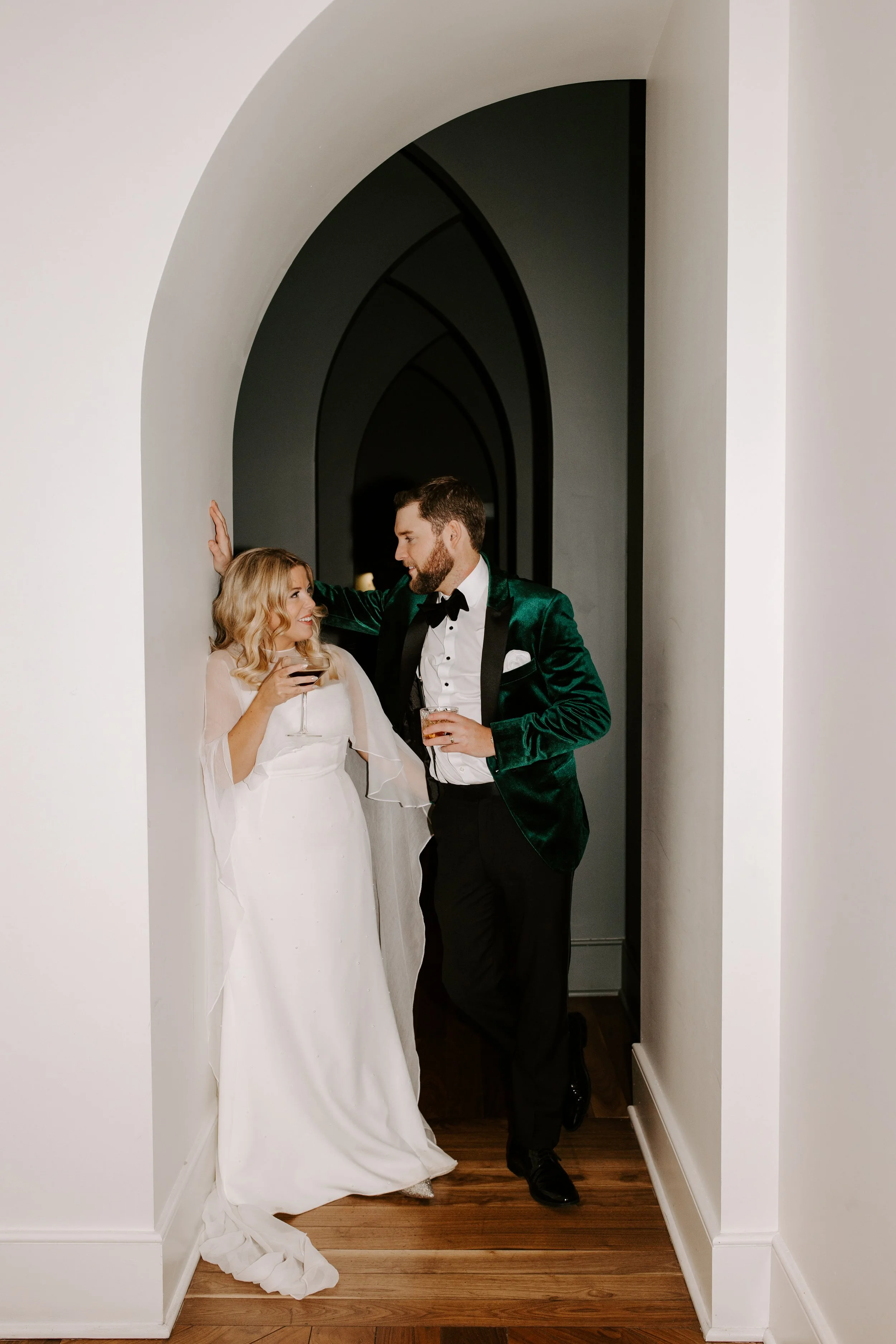A bride and groom in tuxedo and wedding gown, talking and holding drinks, standing in a hallway with arched doorways.